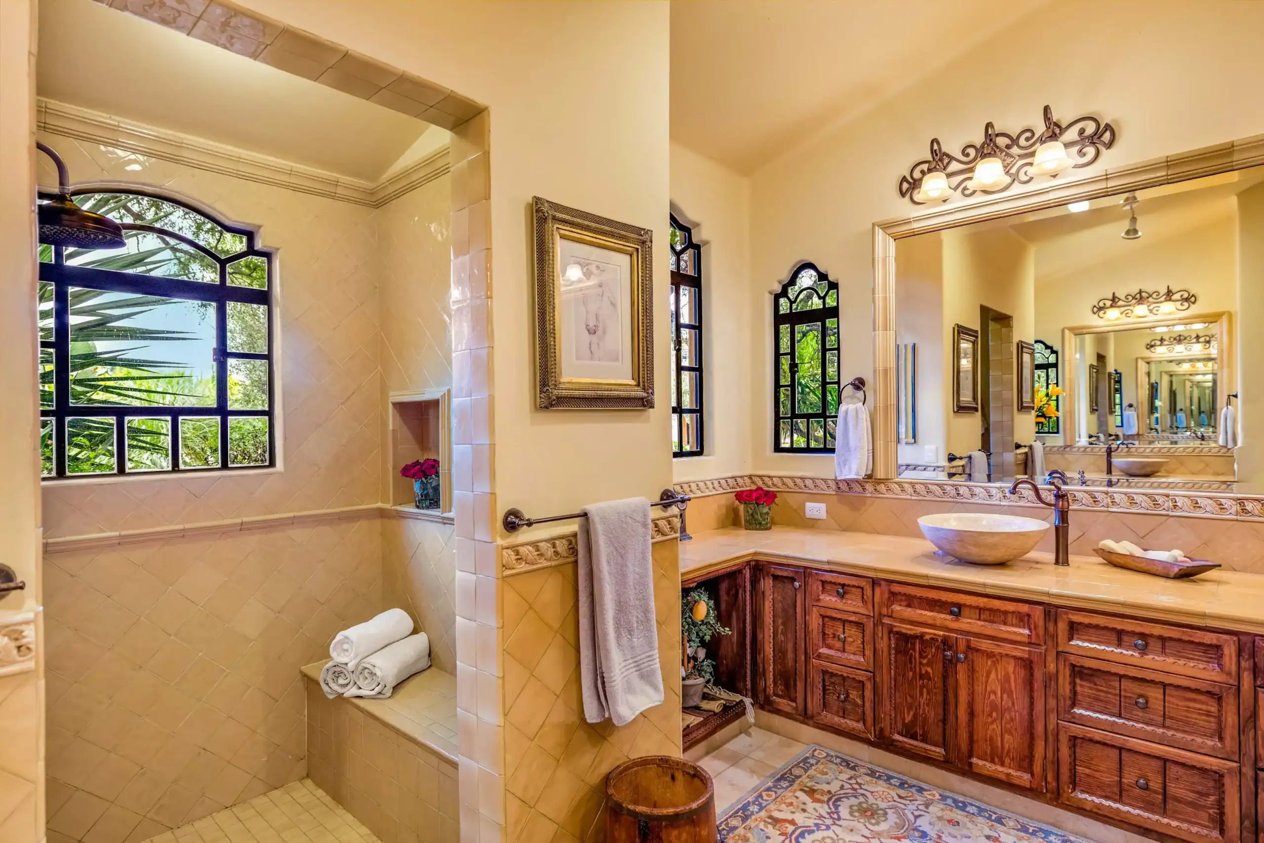 Elegant bathroom with beige tile, wood vanity, and dual sinks, featuring arched windows and a tiled shower area with greenery outside, San Miguel de Allende