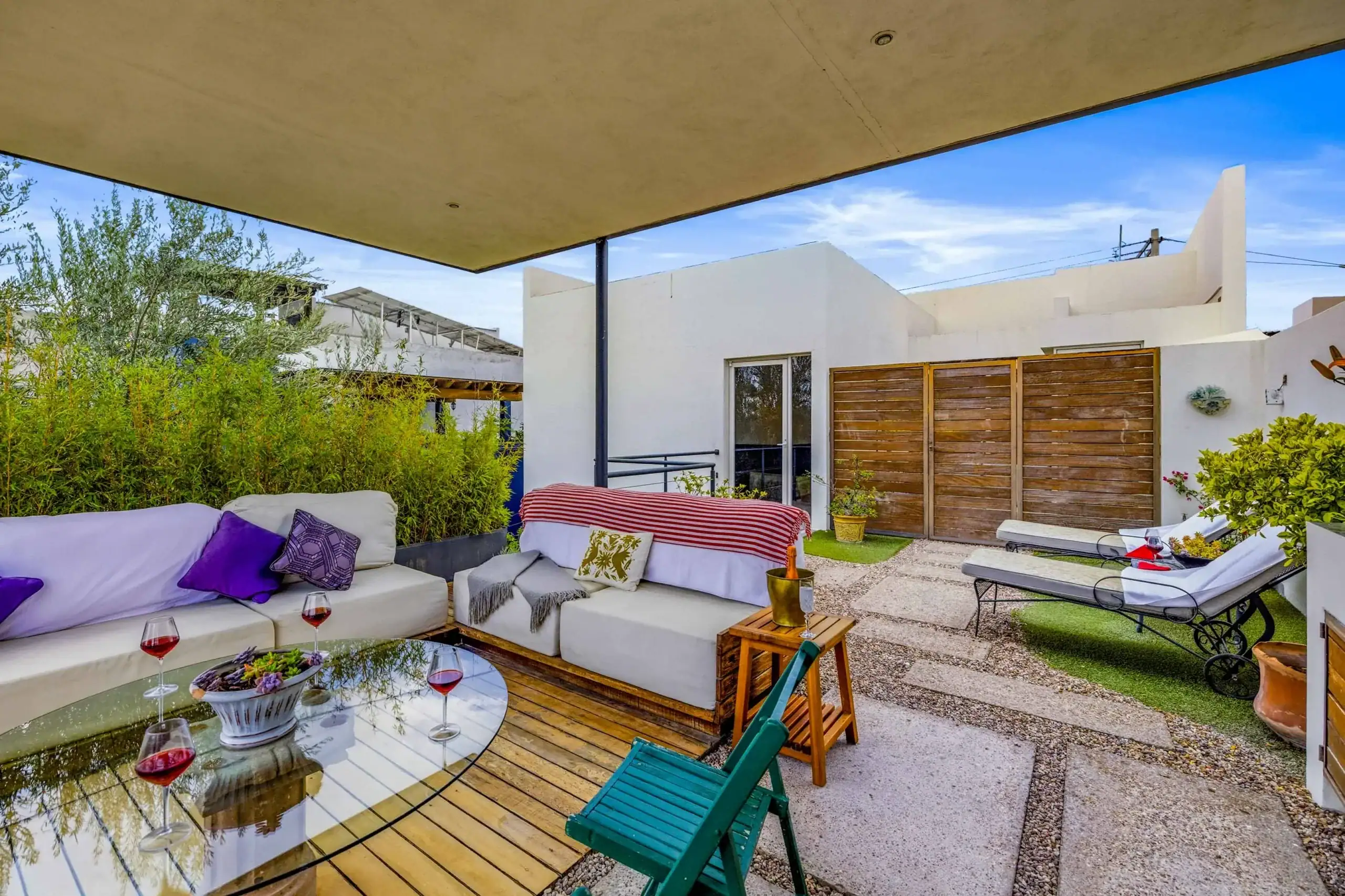 Outdoor patio seating area with a white sectional sofa, purple cushions, and a glass coffee table with red wine glasses, surrounded by greenery behind a beige canopy, San Miguel de Allende