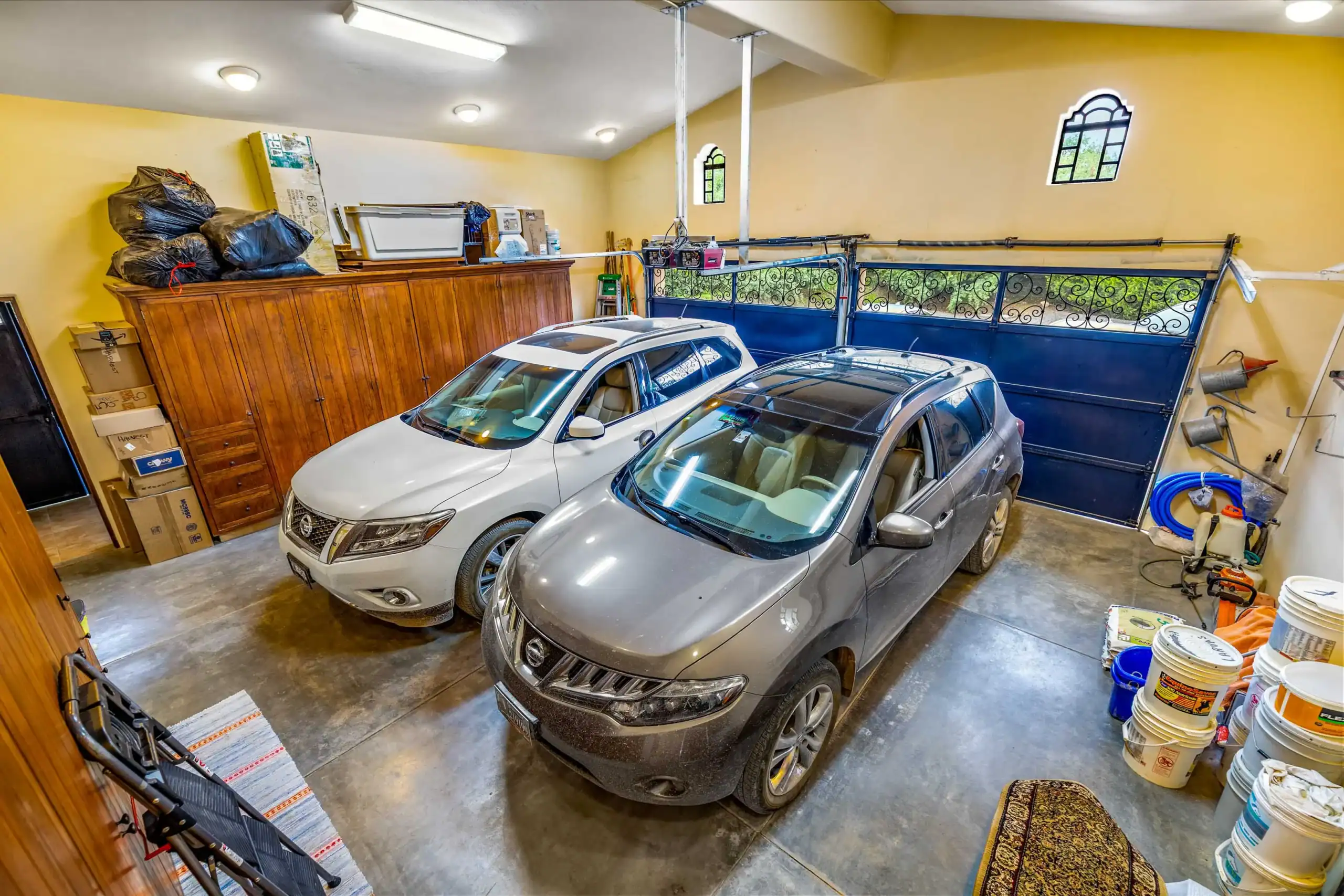 Two silver SUVs parked in a home garage with yellow walls, wooden cabinets, and storage boxes along the left side, San Miguel de Allende