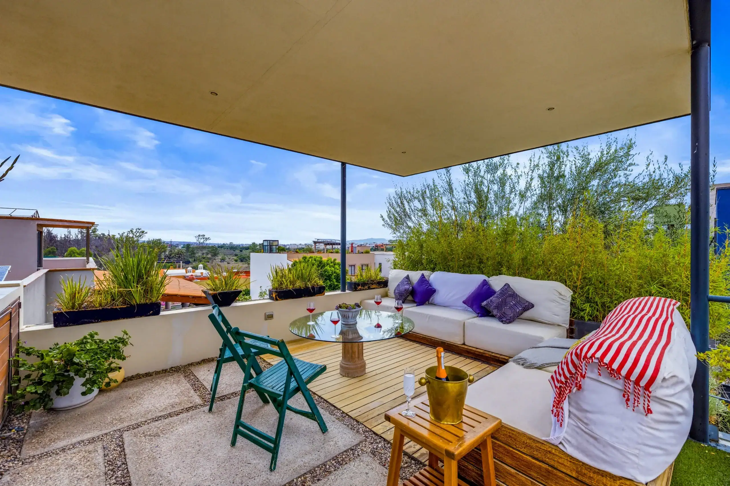 Outdoor rooftop patio with a beige shade sail, white sectional sofa with purple pillows, glass round table, and wine glasses on a sunny day, San Miguel de Allende