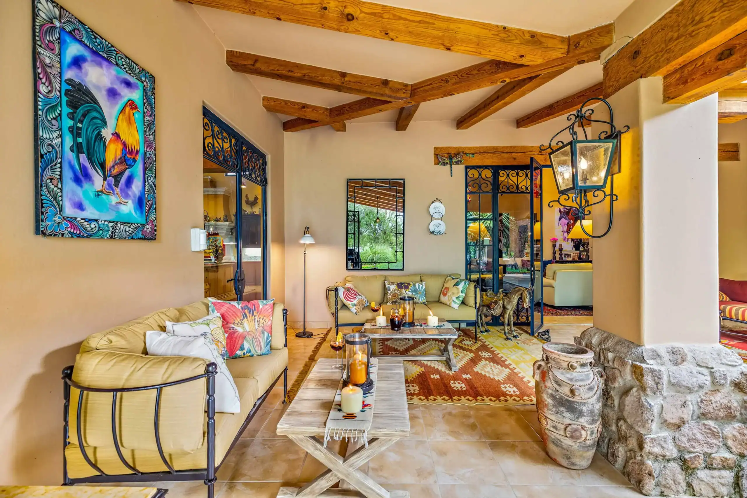 Sunlit living room with beige couches and colorful cushions, a large vibrant rooster painting on the left wall, and exposed wooden ceiling beams, San Miguel de Allende