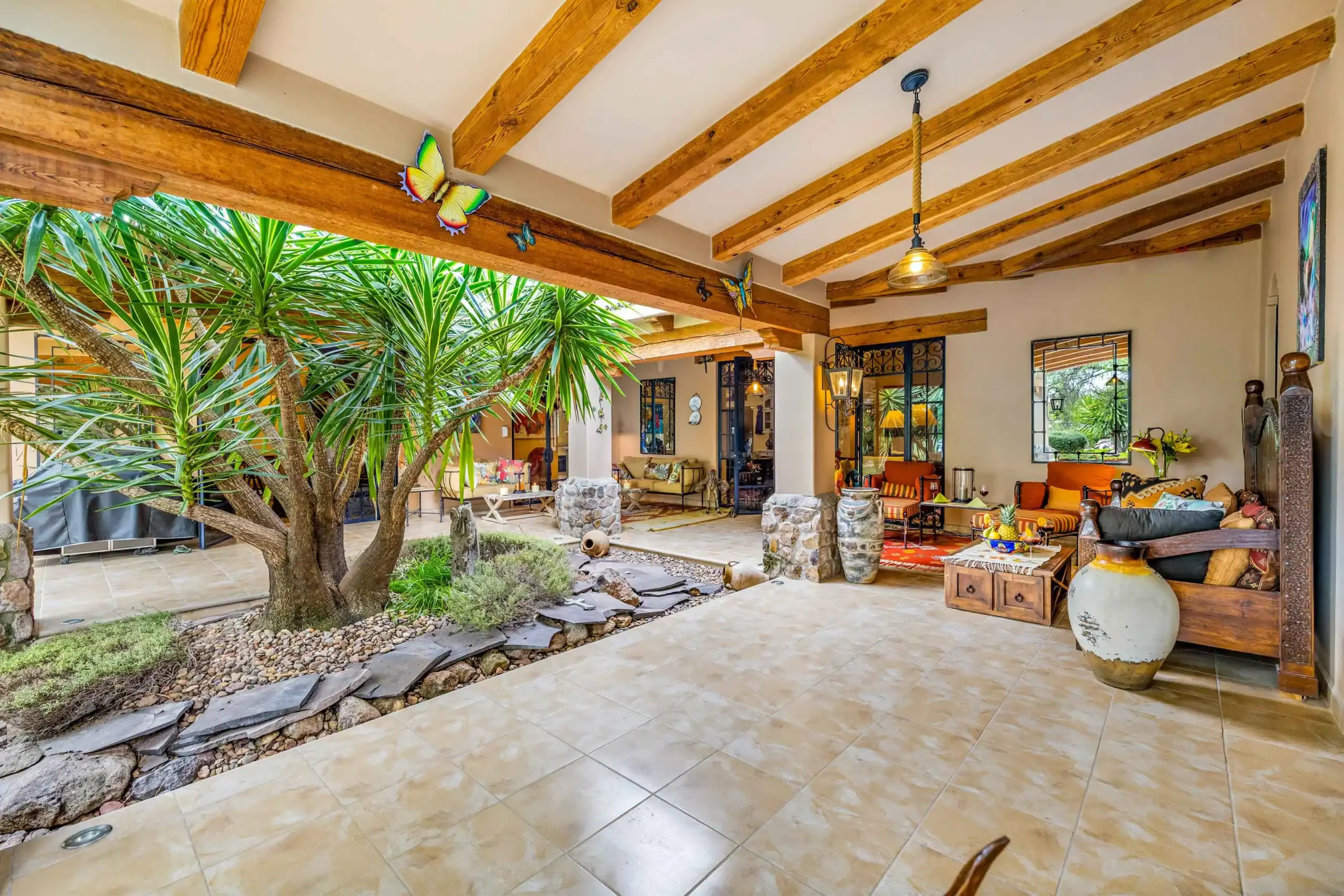 Covered patio with wooden ceiling beams, lush indoor plants, and colorful butterfly ornaments on the beam. A seating area with cushions visible in the background, San Miguel de Allende