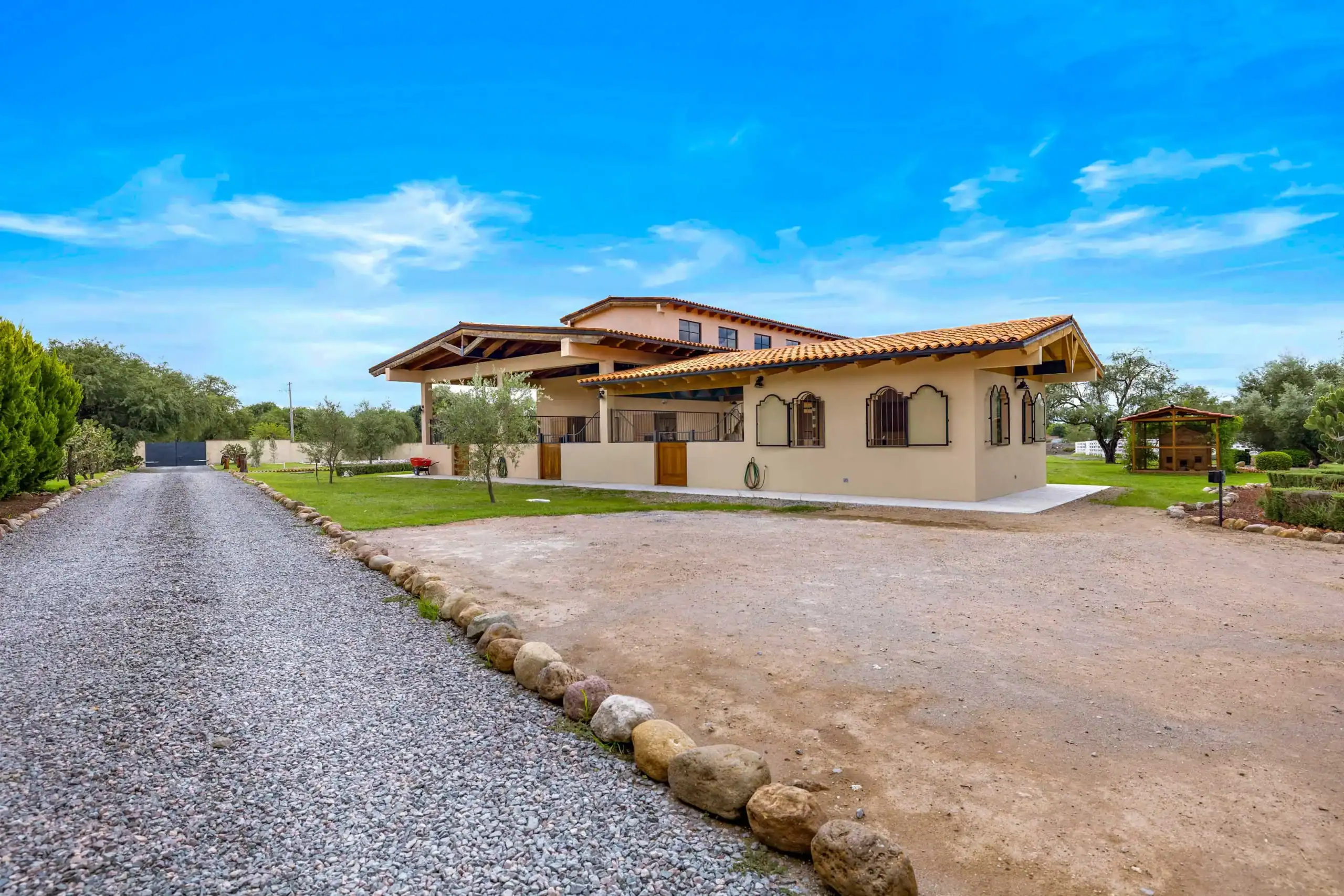 Beige single-story house with orange terracotta roof, surrounded by a gravel driveway and green lawn under a bright blue sky, San Miguel de Allende