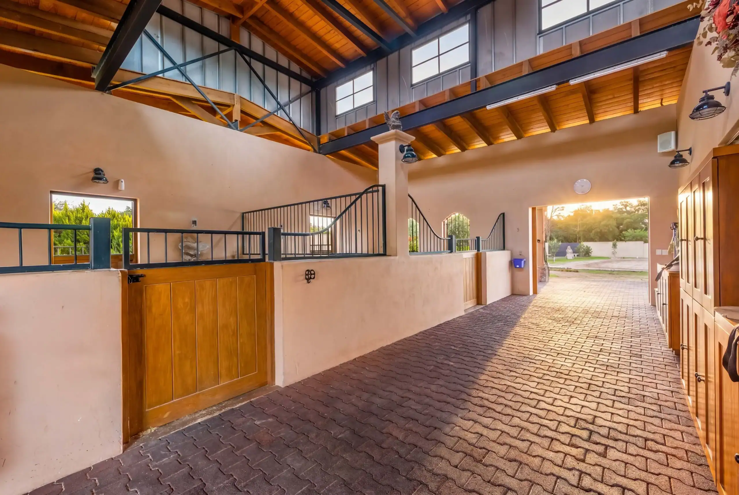 Sunlit horse stable interior with beige walls, wooden stalls, and a brick-paved central aisle, San Miguel de Allende