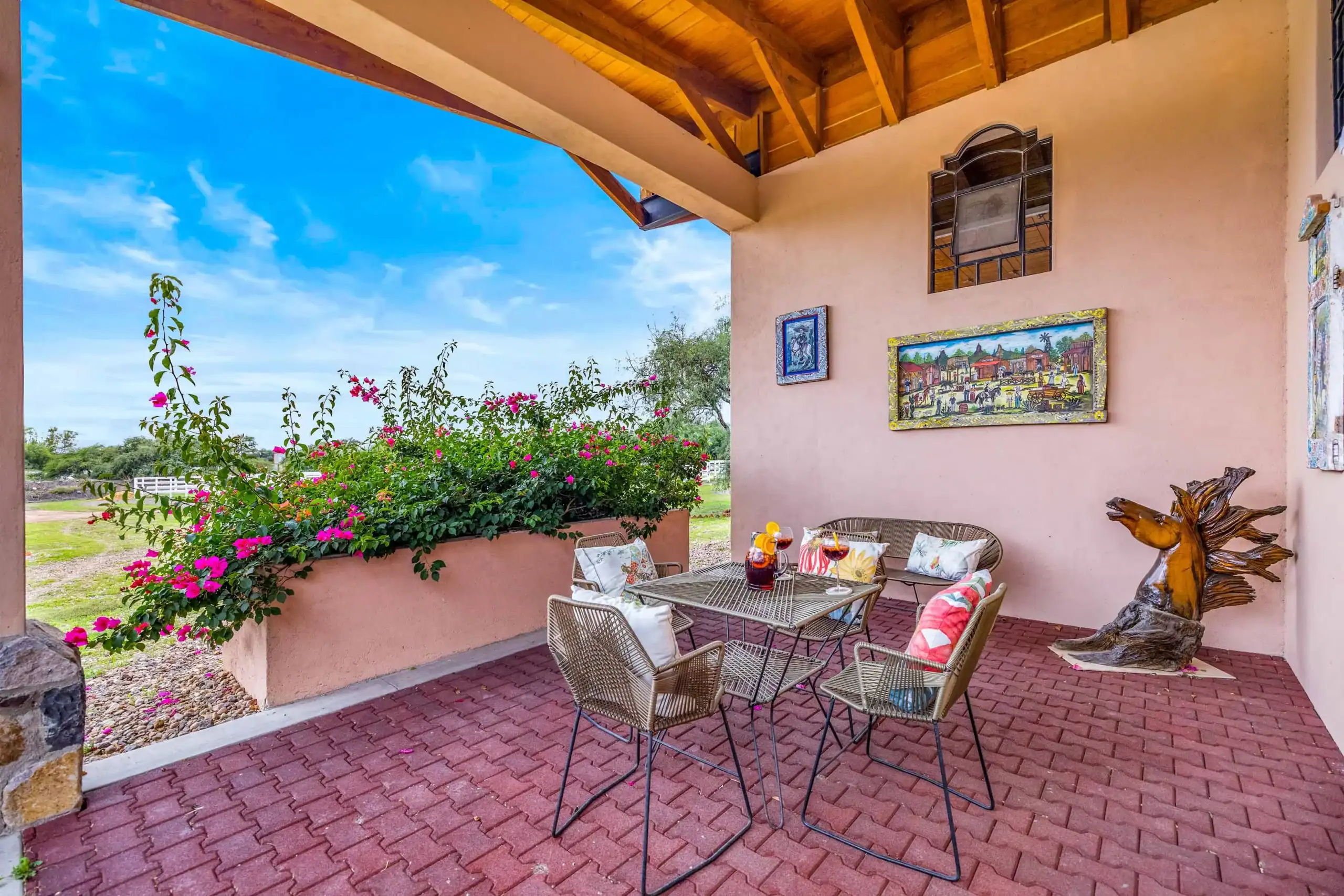 Outdoor patio with pink stucco walls, wicker chairs around a metal table with drinks, flowering shrubs, and wall art under a wooden pergola, San Miguel de Allende