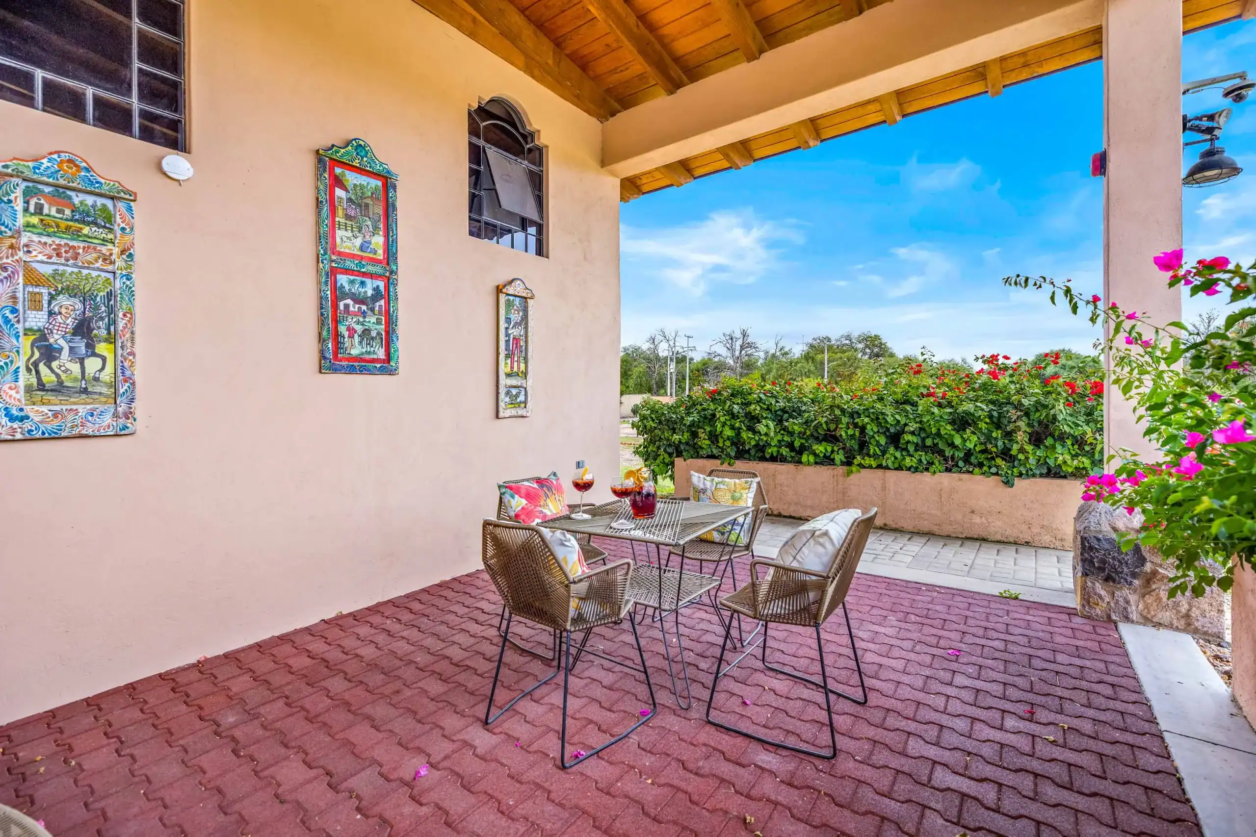 Outdoor patio with pink stucco wall, colorful tiled panels, and wicker chairs around a metal table with drinks under a wooden pergola, blue sky over greenery, San Miguel de Allende