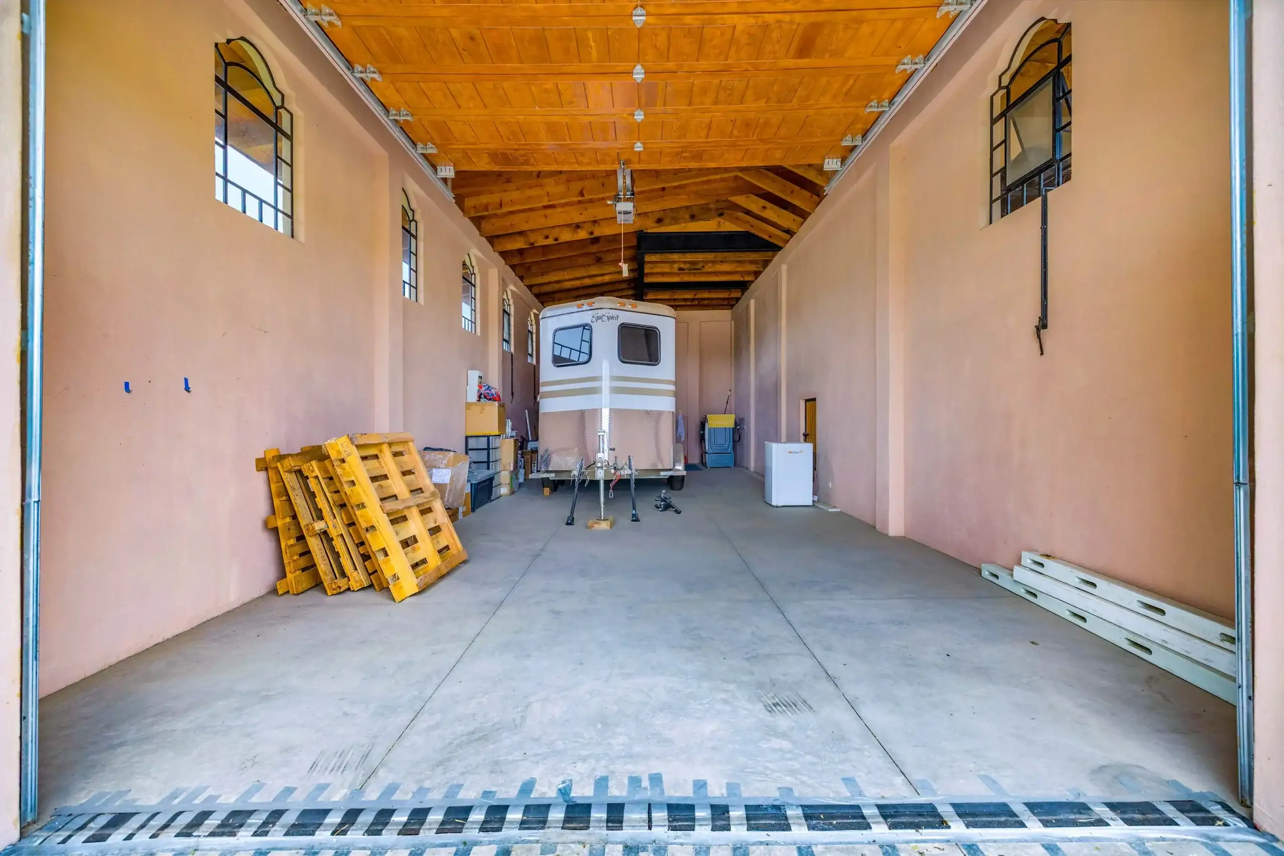 Interior of a large storage shed with a travel trailer parked in the center, pallets on the left and windows along peach walls, San Miguel de Allende