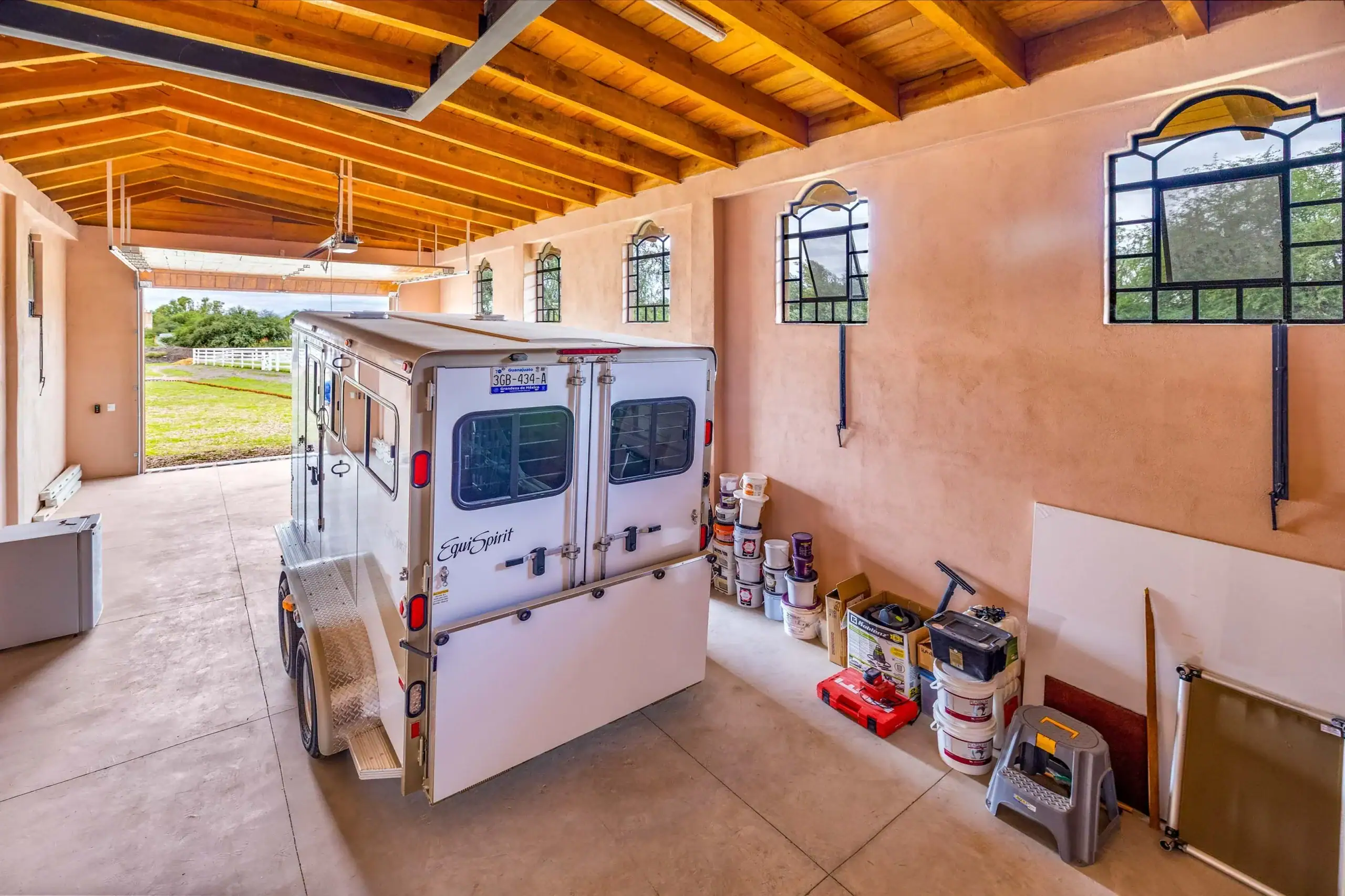 White horse trailer parked inside a garage with a wooden beam ceiling and storage buckets along the wall; open door shows a grassy yard outside, San Miguel de Allende