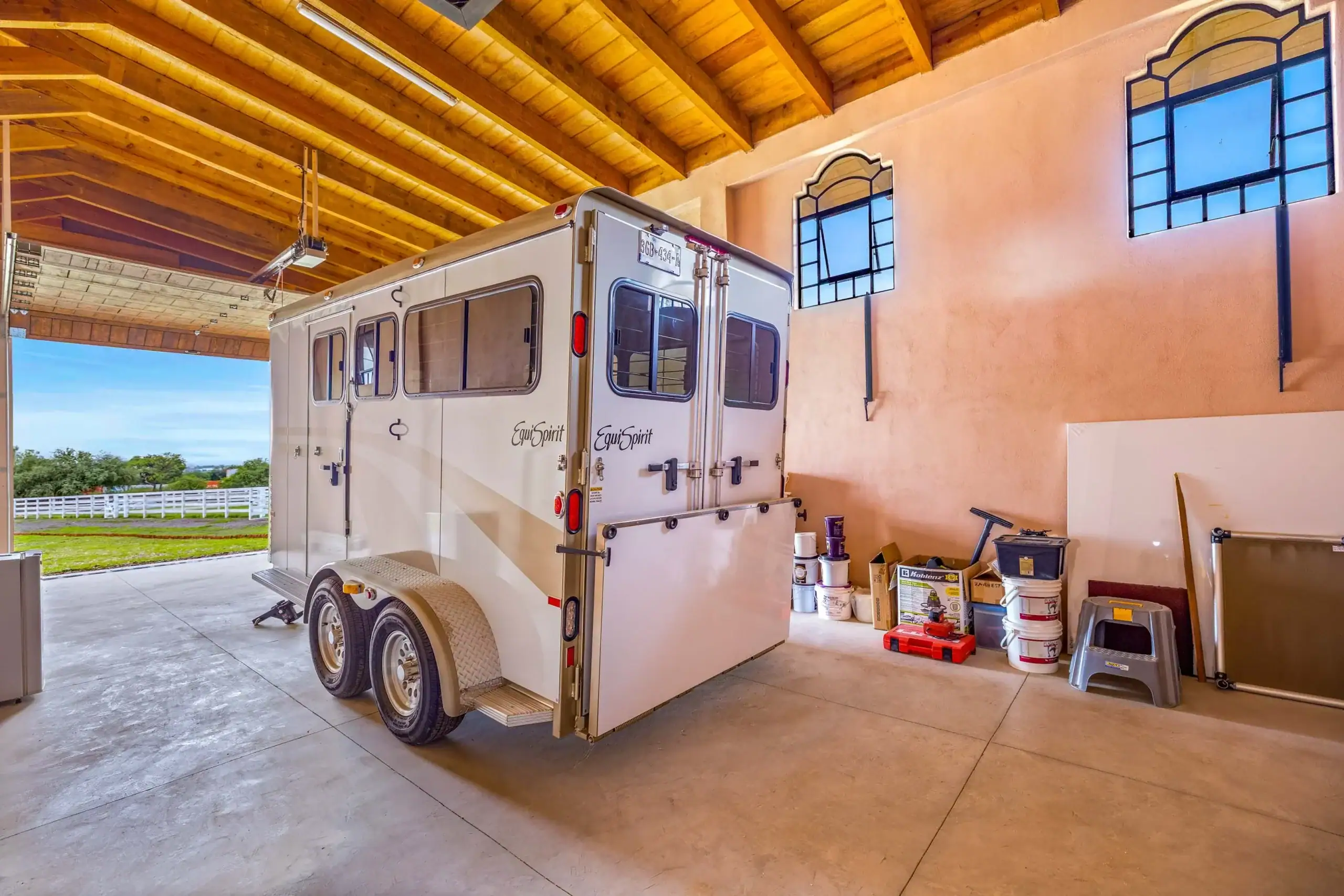 A silver horse trailer inside a garage with a wooden beam ceiling, windows on the trailer, and various tools and paint cans stacked against the wall nearby, San Miguel de Allende