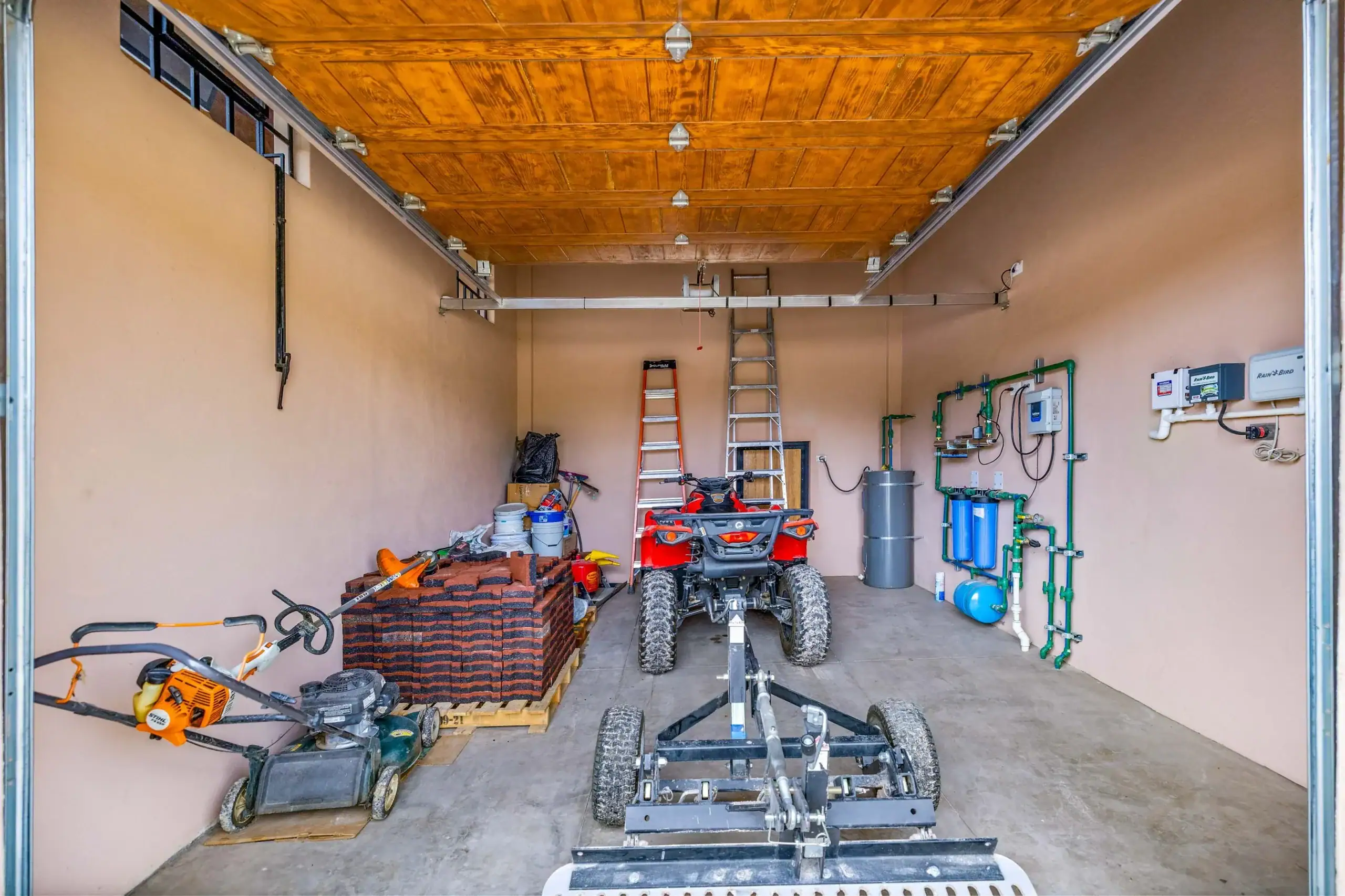 A spacious garage filled with tools and equipment: ladders, stacked bricks, a ride-on quad bike, and wall-mounted pipes along beige walls under a wooden ceiling, San Miguel de Allende