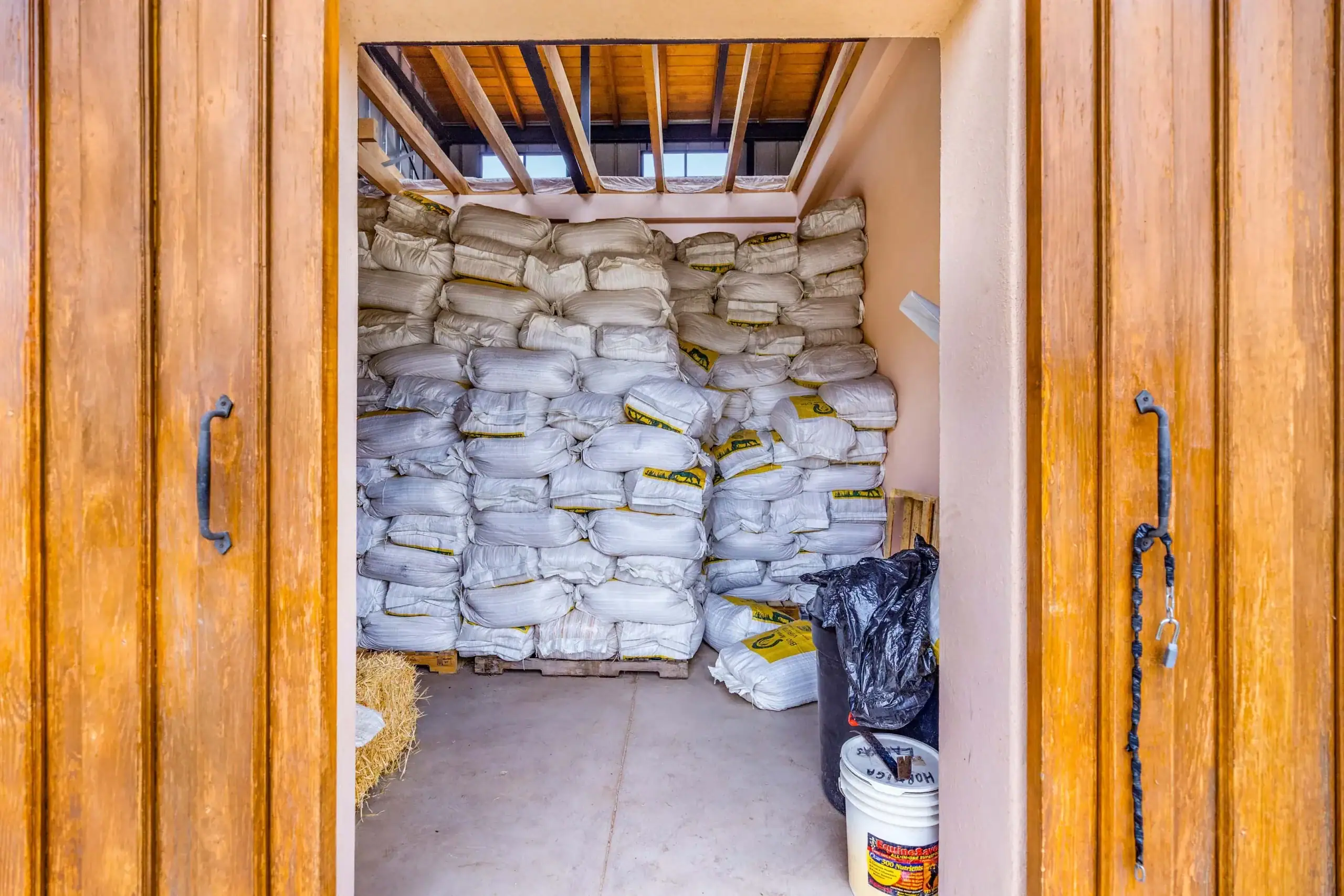 Open wooden doors reveal a storage room packed with stacked white bags, with hay bales on the left and a black trash bag on the floor to the right, San Miguel de Allende