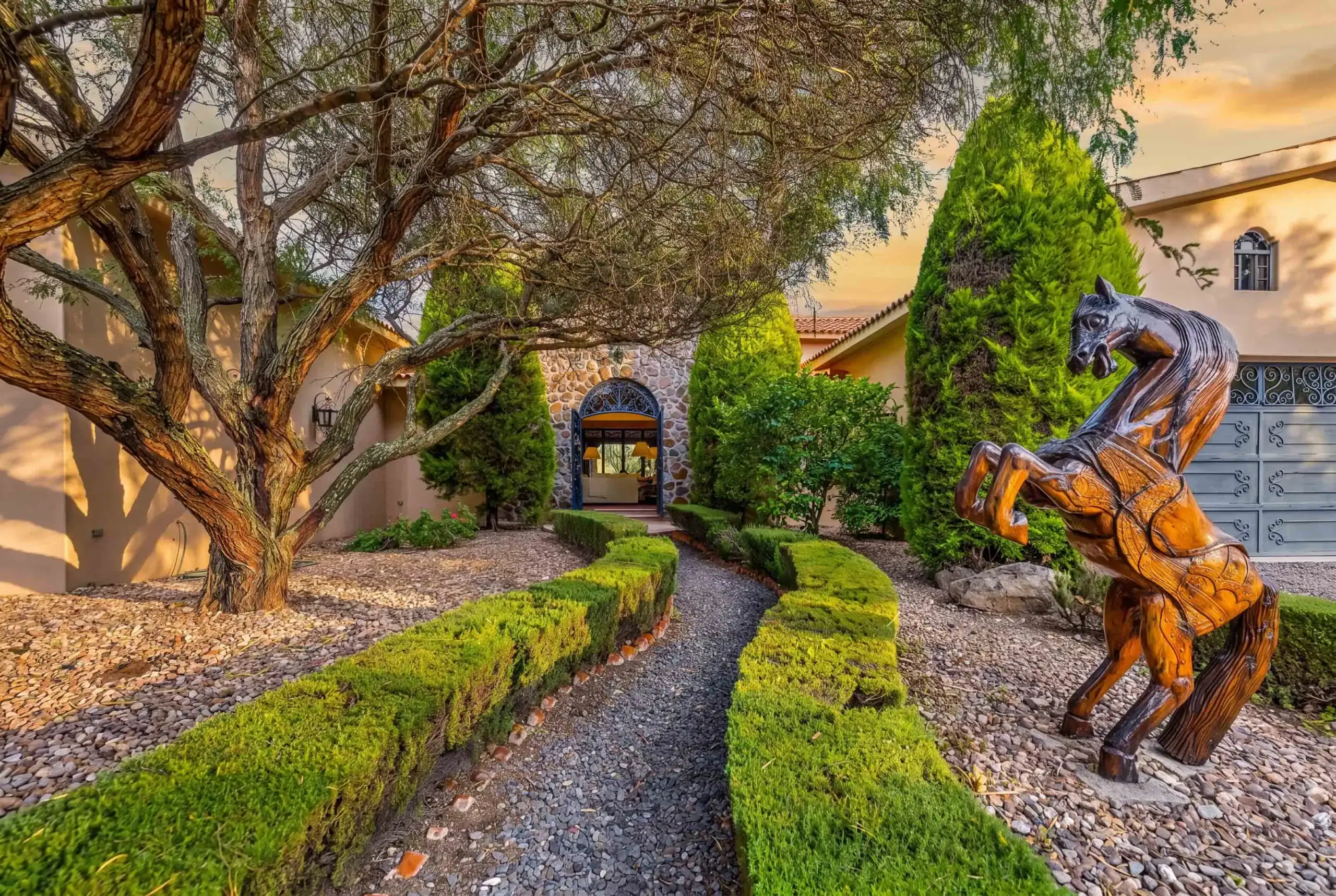 Wooden horse statue rearing in a Mediterranean courtyard with trimmed hedges, a stone arch doorway, and a large tree overhead, San Miguel de Allende