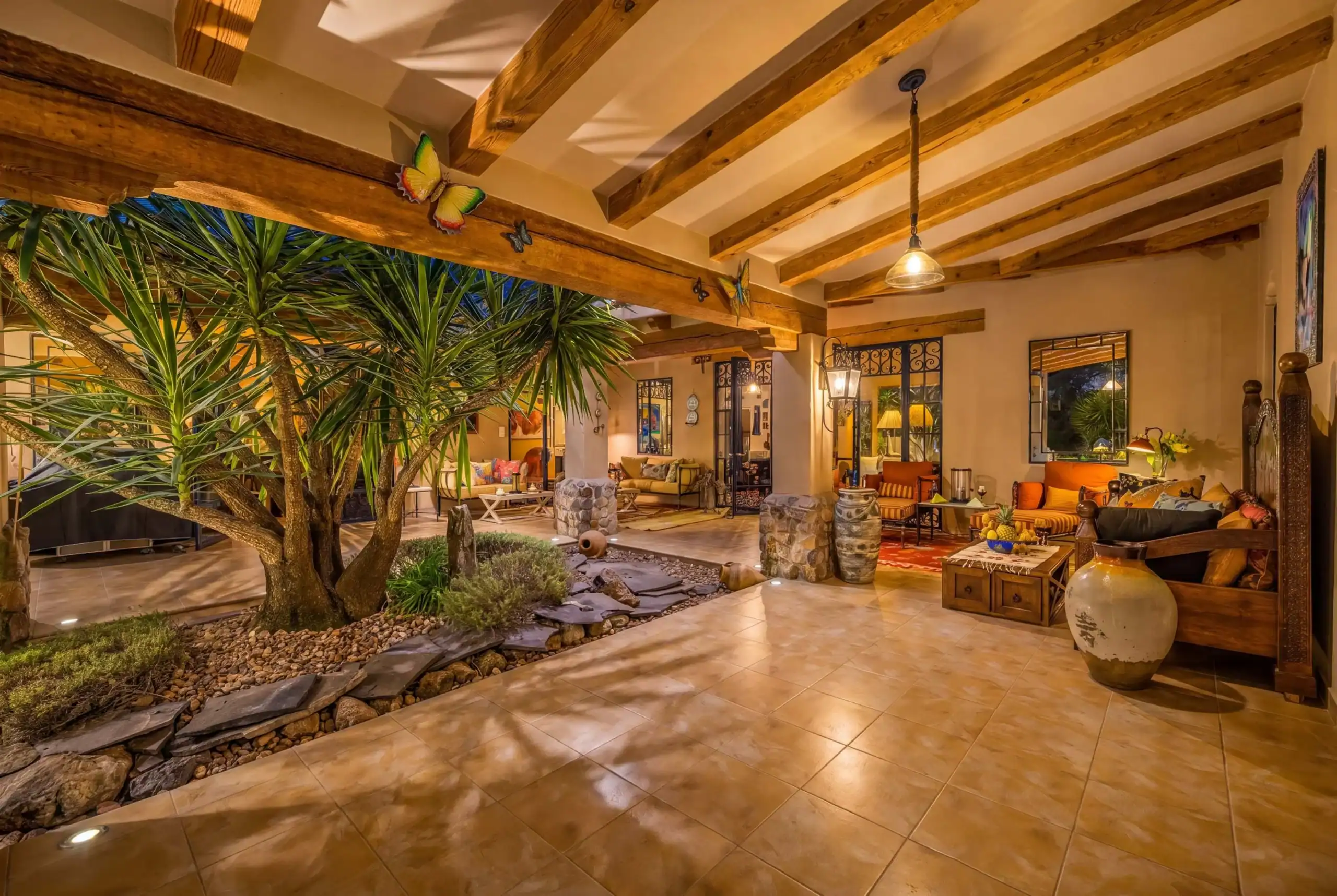 Indoor courtyard living space with exposed wooden beams, warm pendant light, and a seating area of orange cushions and sofas surrounded by tropical plants, San Miguel de Allende