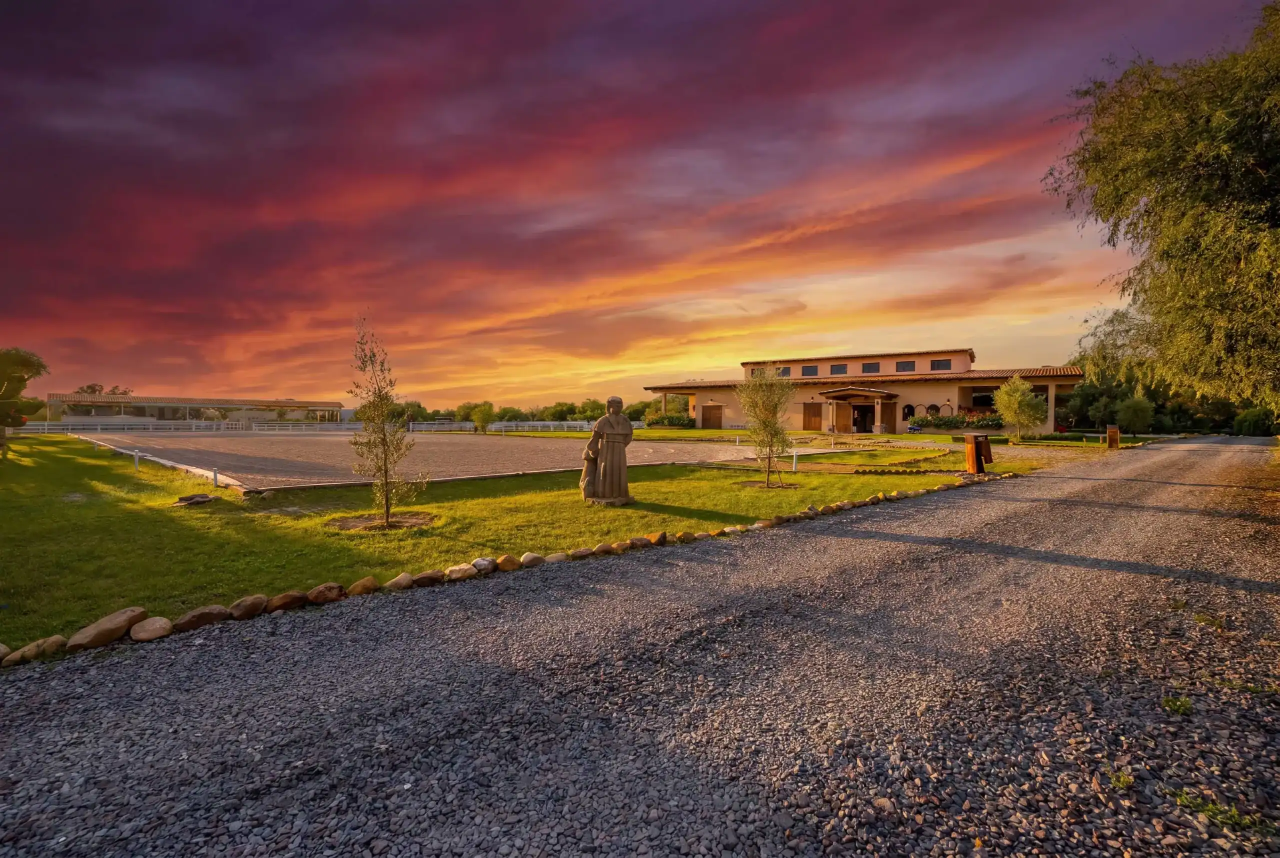 Sunset over a rural campus-like area with a statue in the foreground, gravel paths, green lawn, and a beige building on the right, San Miguel de Allende