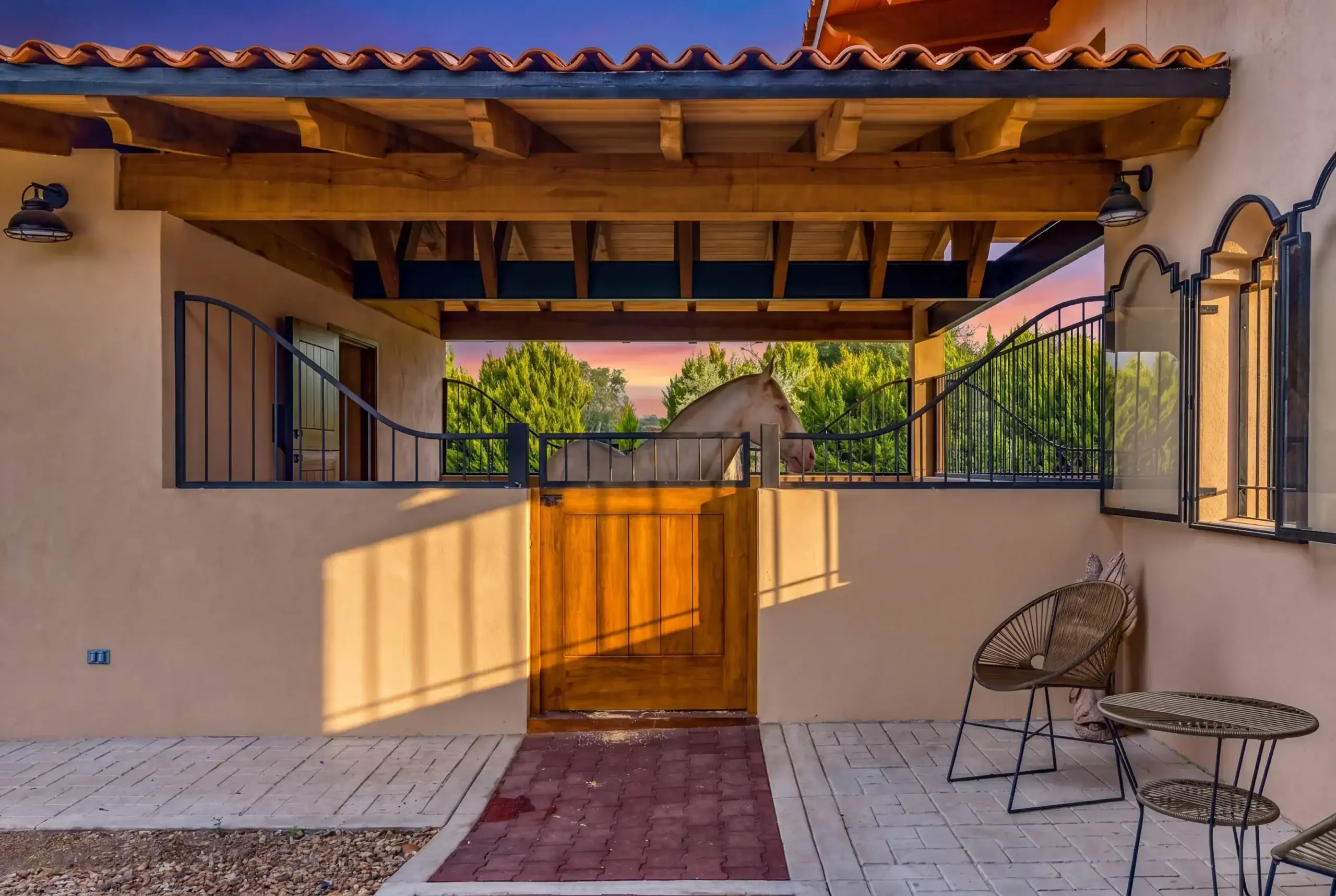 Sunlit courtyard with stucco walls, a wooden pergola, and a white horse inside a black metal gate; patio chairs on the right, San Miguel de Allende