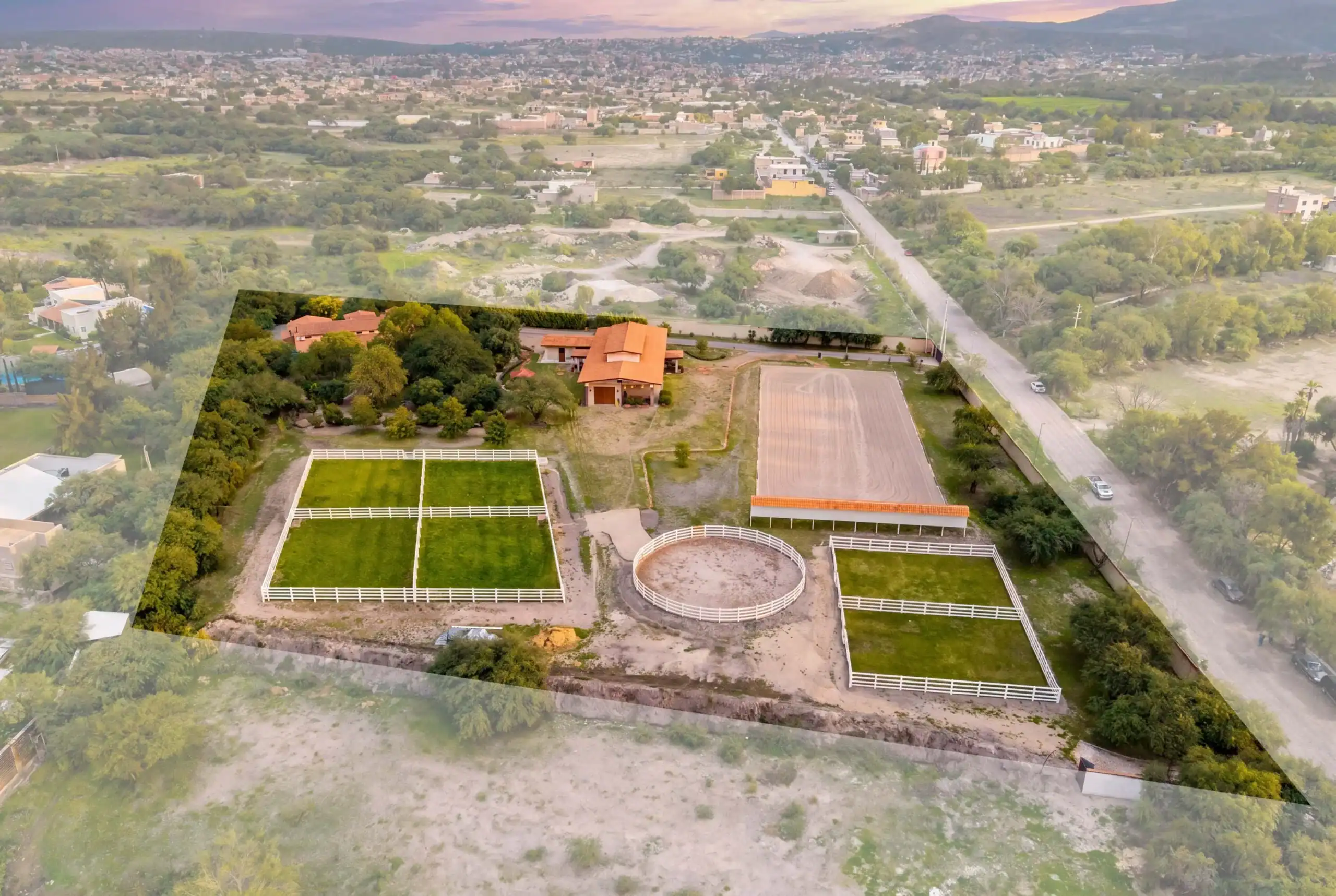 Aerial view of a property featuring a circular riding arena, a orange-roofed barn, fenced paddocks, and surrounding green fields and trees in a suburban area, San Miguel de Allende