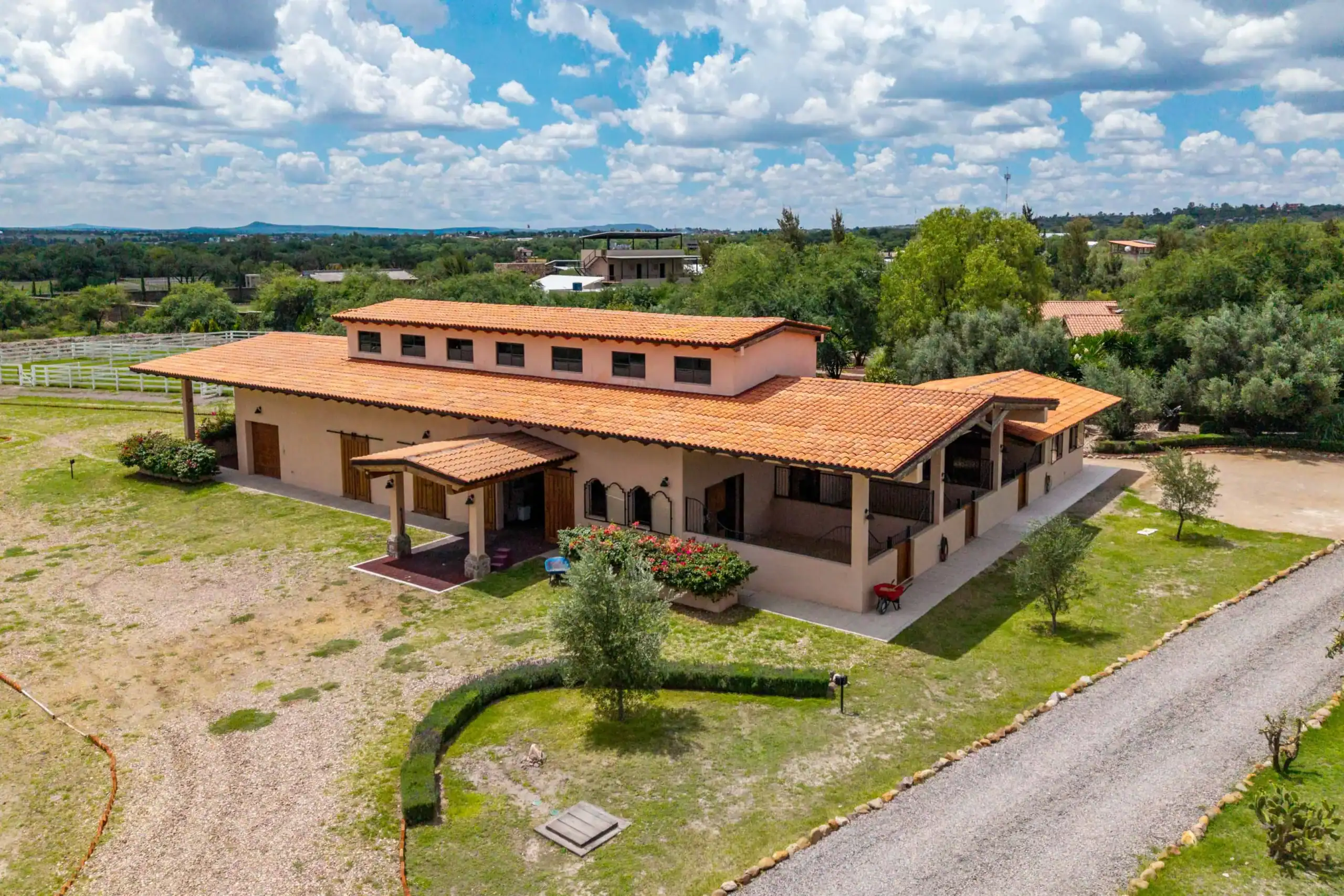 Ranch-style building with orange-tiled roof in a spacious rural yard, gravel paths, and green trees under a cloudy sky, San Miguel de Allende