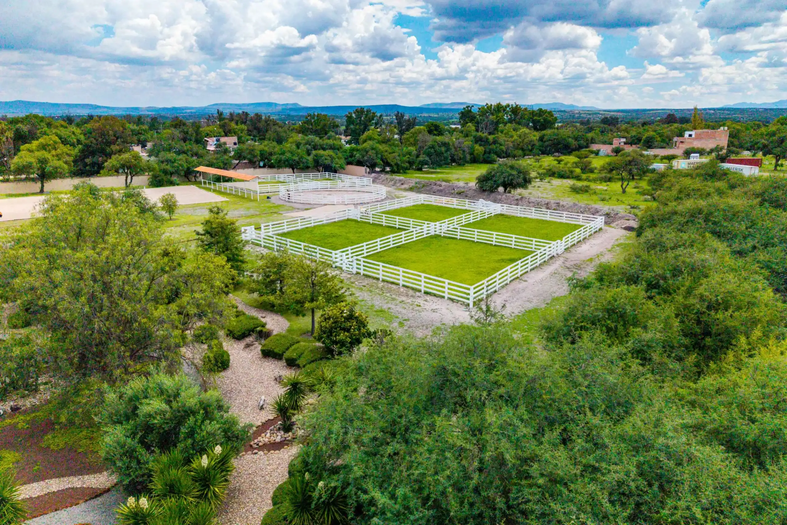 Aerial view of a ranch with white fences enclosing grassy paddocks, a circular arena, and a tan-roofed shelter among trees under a cloudy blue sky, San Miguel de Allende
