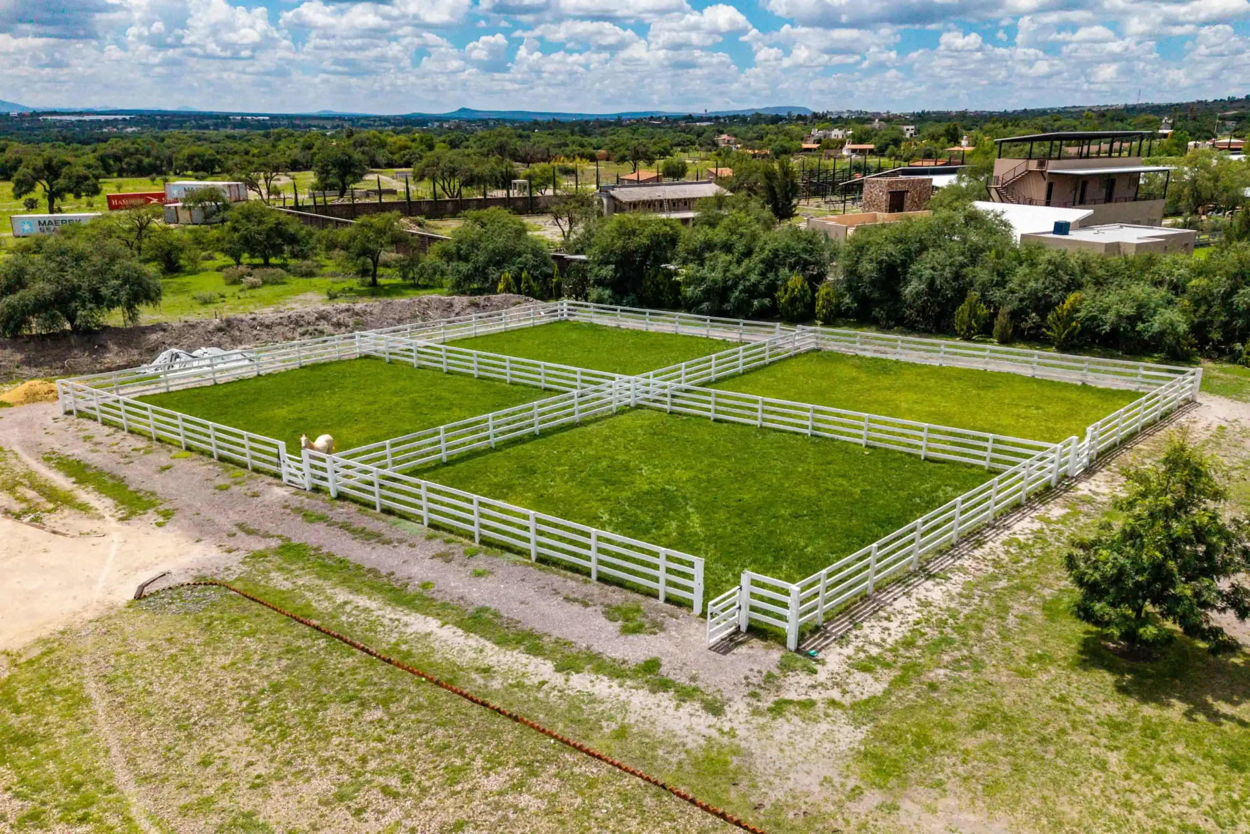 Aerial view of white-fenced horse paddocks with green grass and distant buildings under a cloudy blue sky, San Miguel de Allende