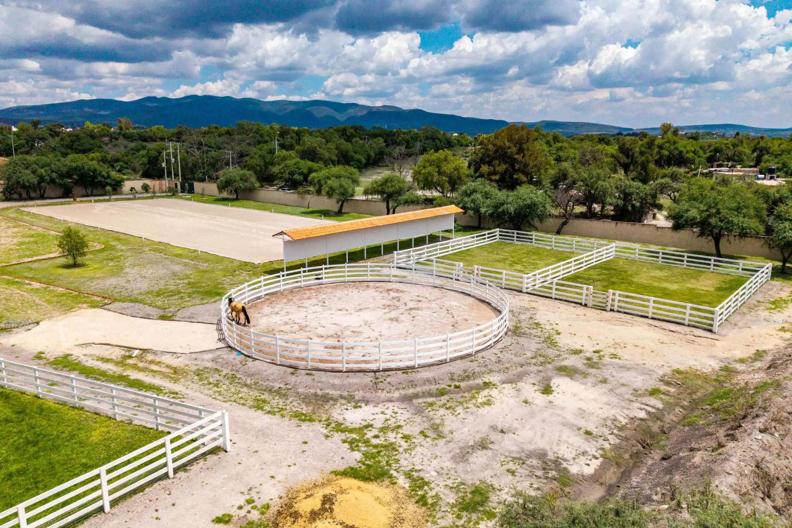Round riding arena with a white fence and a single horse, set in grassy paddocks and distant hills under a cloudy sky, San Miguel de Allende