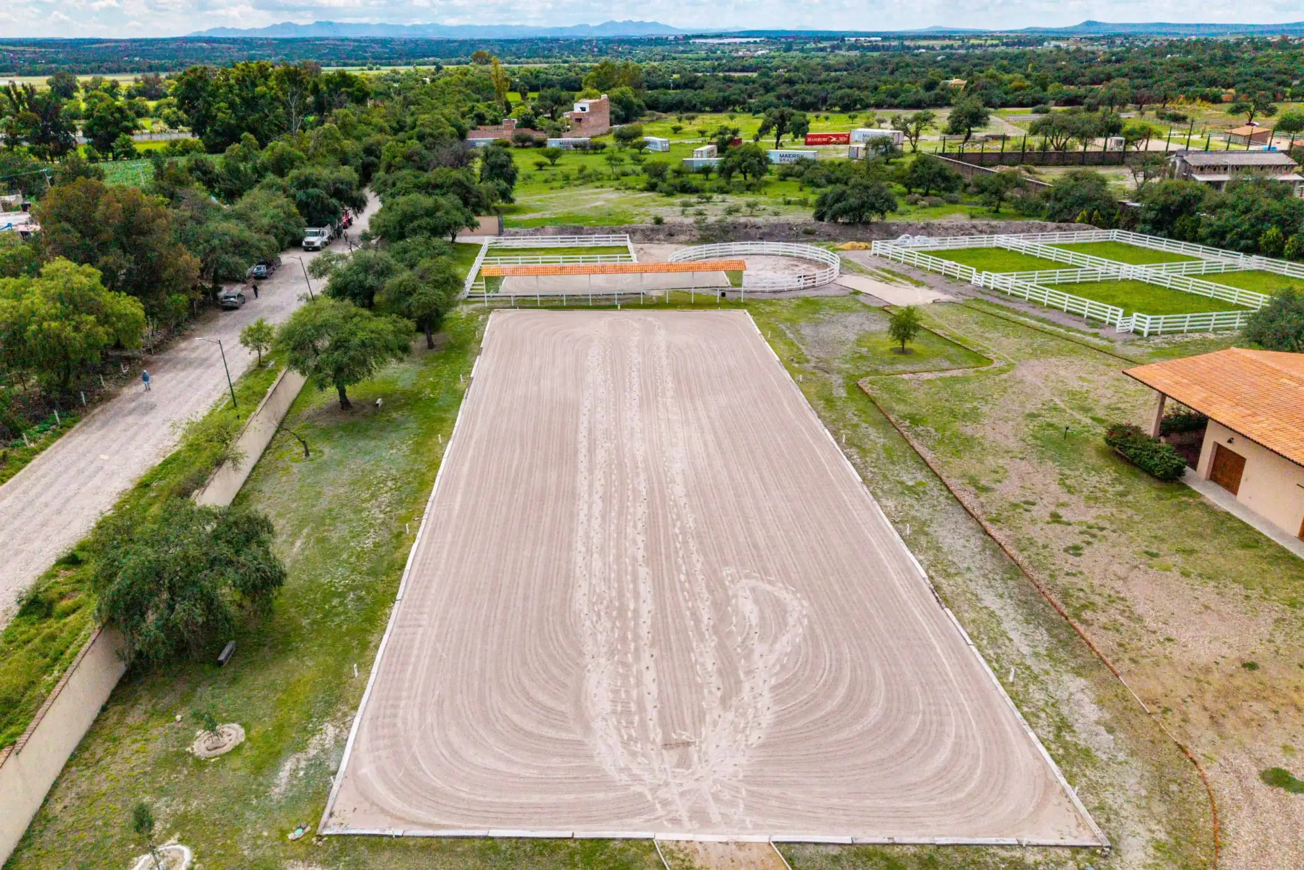 Aerial view of an equestrian facility with a large rectangular riding arena, a circular pen, and white fences amid green fields and trees, San Miguel de Allende
