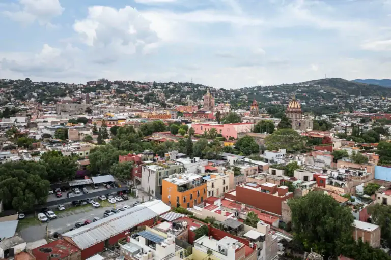 Panoramic view of a colorful hillside city with a prominent orange-domed church and a cloudy sky backdrop, San Miguel de Allende