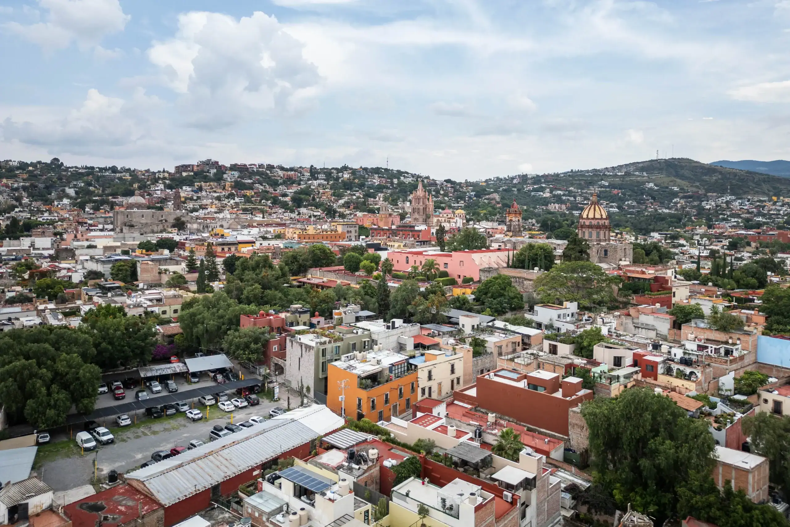 Panoramic view of a colorful hillside city with a prominent orange-domed church and a cloudy sky backdrop, San Miguel de Allende