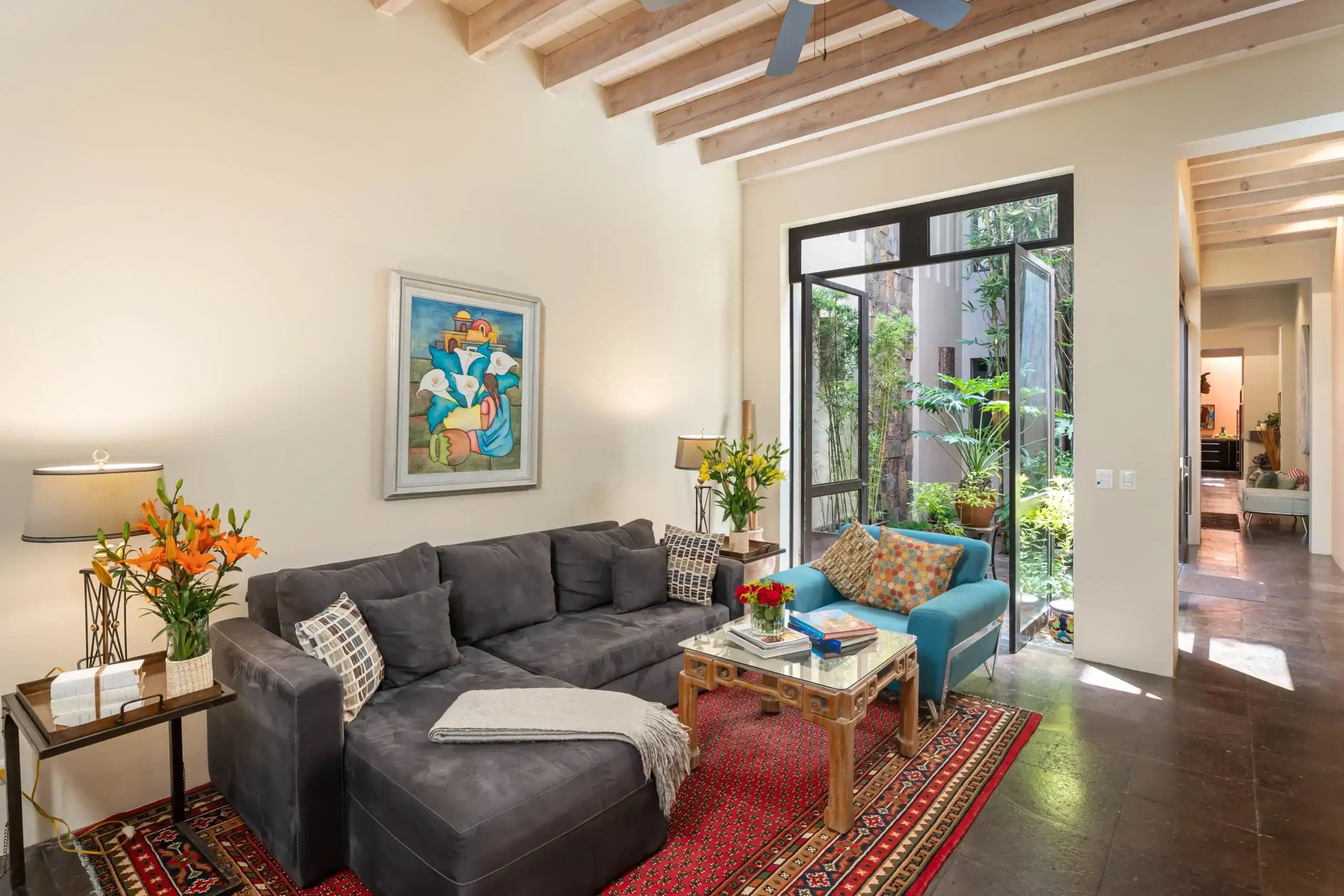 Bright living room with a dark gray sectional sofa, colorful cushions, and glass coffee table; large glass doors open to a courtyard garden with plants, San Miguel de Allende