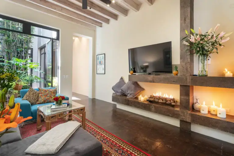 Living room with a large stone fireplace, wall-mounted TV, candles on the hearth, and glass doors opening to greenery; colorful cushions and rug set a cozy mood?, San Miguel de Allende