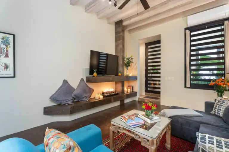 Living room with a stone fireplace, wall-mounted TV, blue patterned cushions, plants, and a glass coffee table under warm lighting, San Miguel de Allende