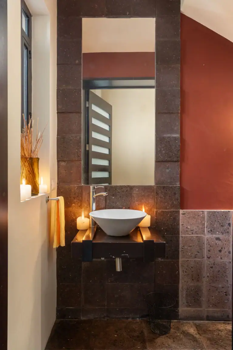 Modern bathroom vanity area with a white vessel sink on a wooden countertop, surrounded by dark stone tiles and lit candles, San Miguel de Allende
