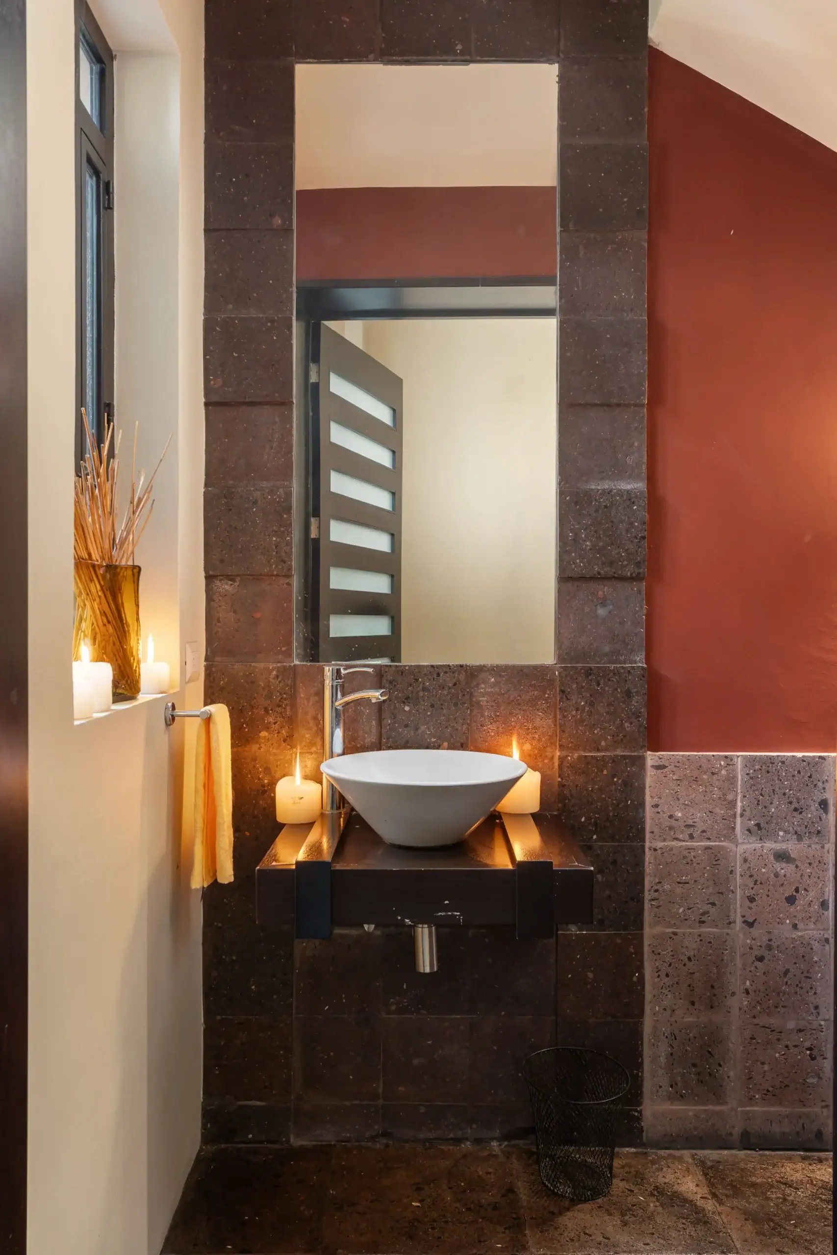 Modern bathroom vanity area with a white vessel sink on a wooden countertop, surrounded by dark stone tiles and lit candles, San Miguel de Allende