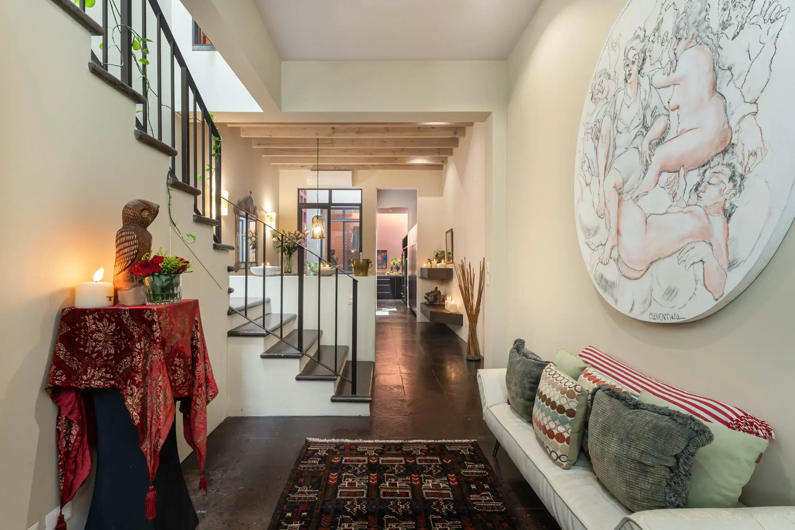 Bright hallway with a black metal staircase on the left, a large circular mural of figures on the right, and a sofa with patterned cushions center-right, San Miguel de Allende