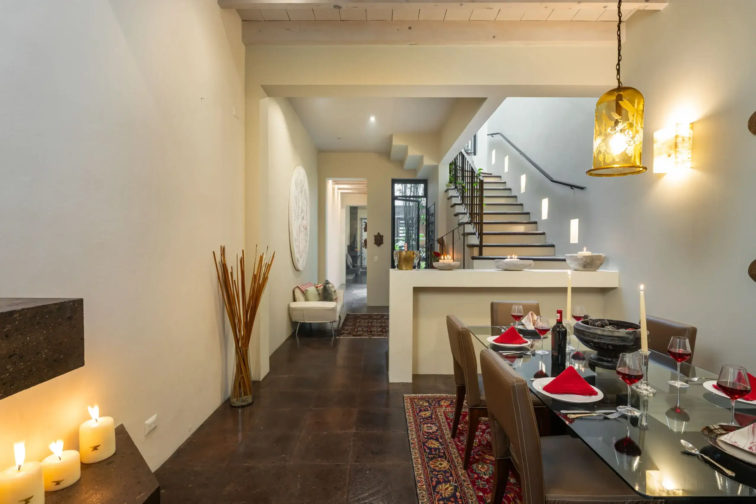 Modern dining area with a glass table set for a meal, red napkins, and candles; staircase visible in the background, San Miguel de Allende