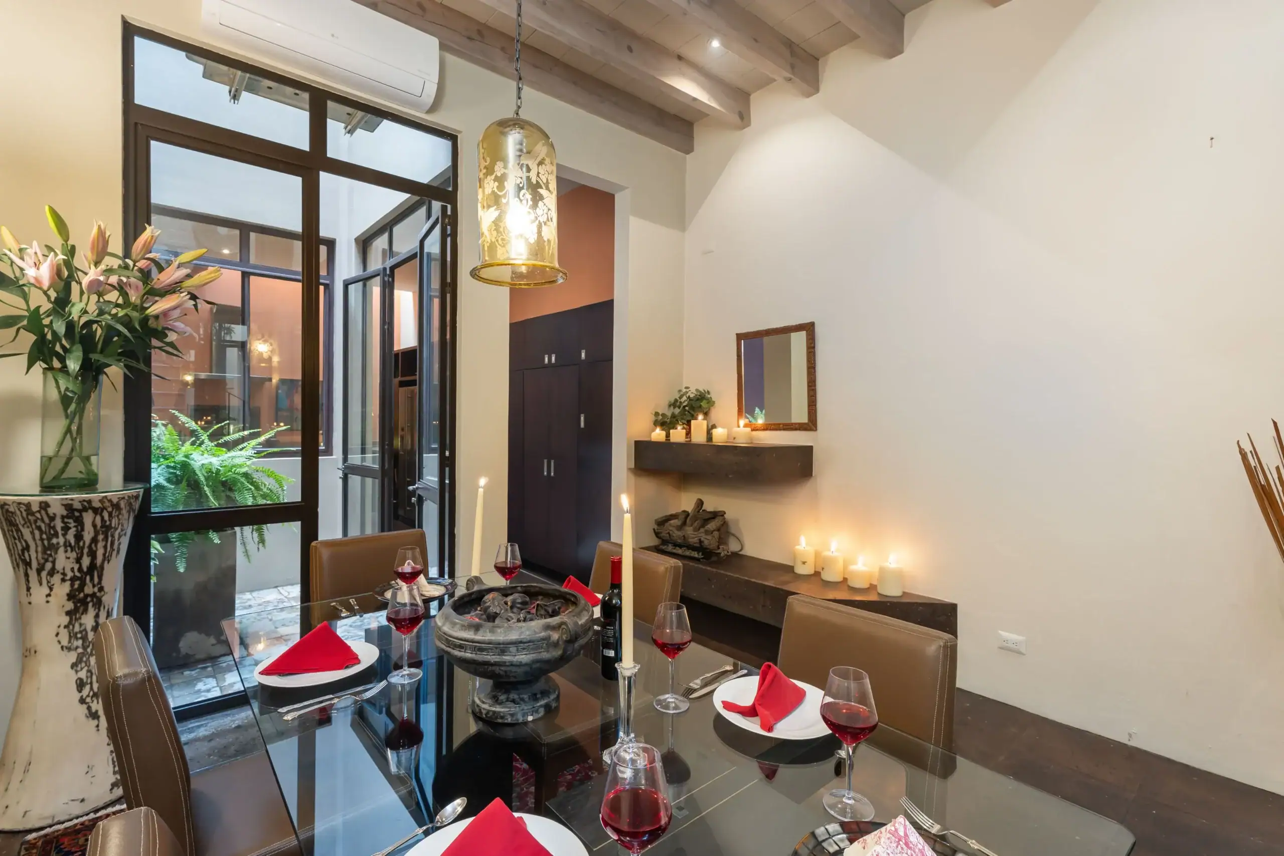 Dining room set for a meal: glass table with red napkins, wine glasses, and a decorative bowl centerpiece in a modern, candlelit space, San Miguel de Allende