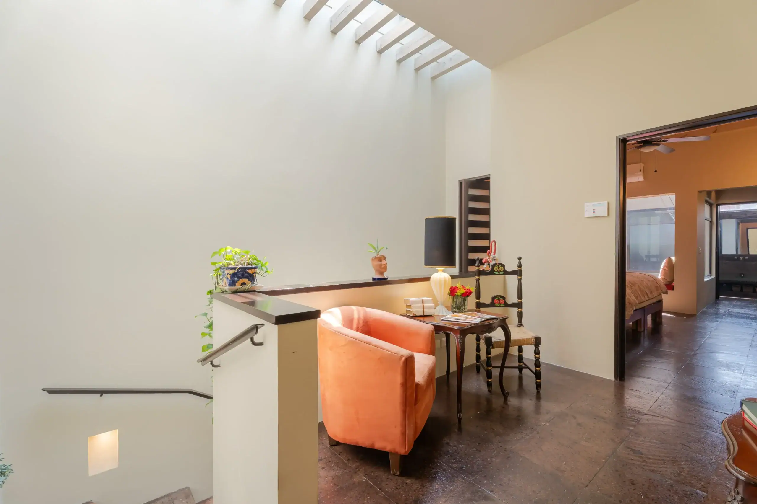 Bright living area with peach chairs at a wooden console, table lamp, and potted plants along a beige wall. A doorway leads to a bedroom visible in the background; skylights overhead, San Miguel de Allende