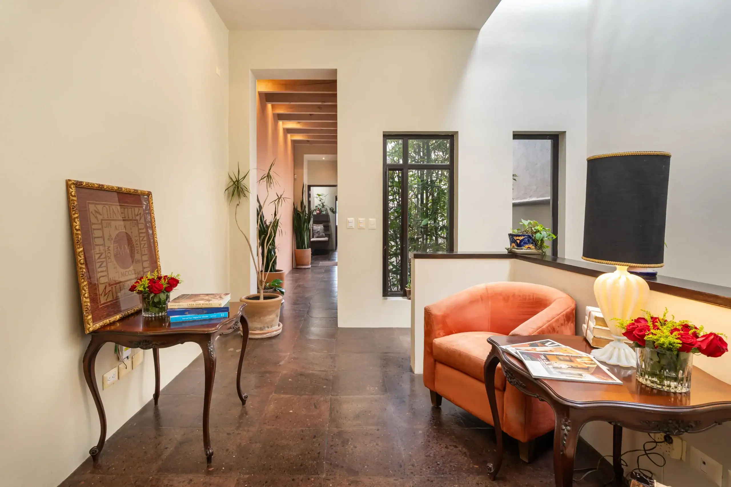 Open living area with orange armchairs, dark wood tables, potted plants, and a hallway with exposed wooden beams in the background, San Miguel de Allende