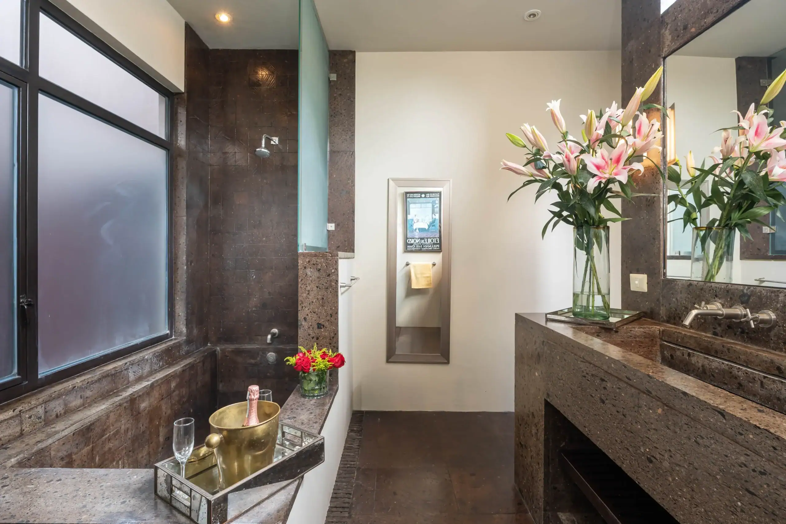 Luxurious bathroom with dark granite surfaces, a glass vase of pink lilies on the counter, and a large mirror reflecting the sink area, San Miguel de Allende