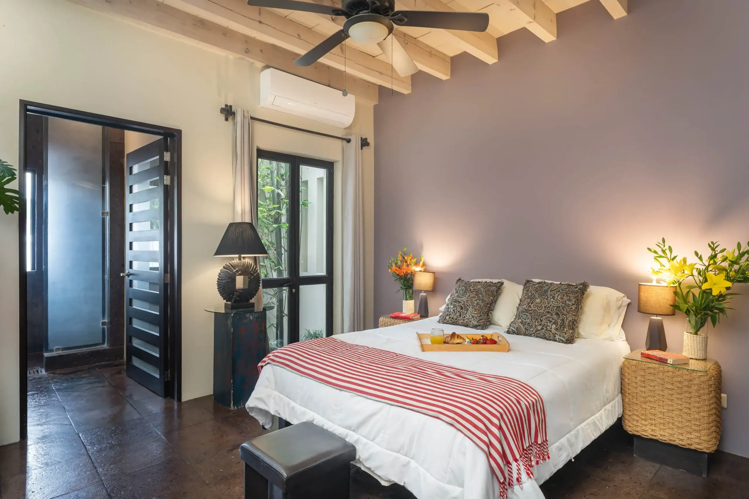 Bright hotel bedroom with white bedding, red-striped throws, and a breakfast tray on the bed; plants and lamps nearby, San Miguel de Allende