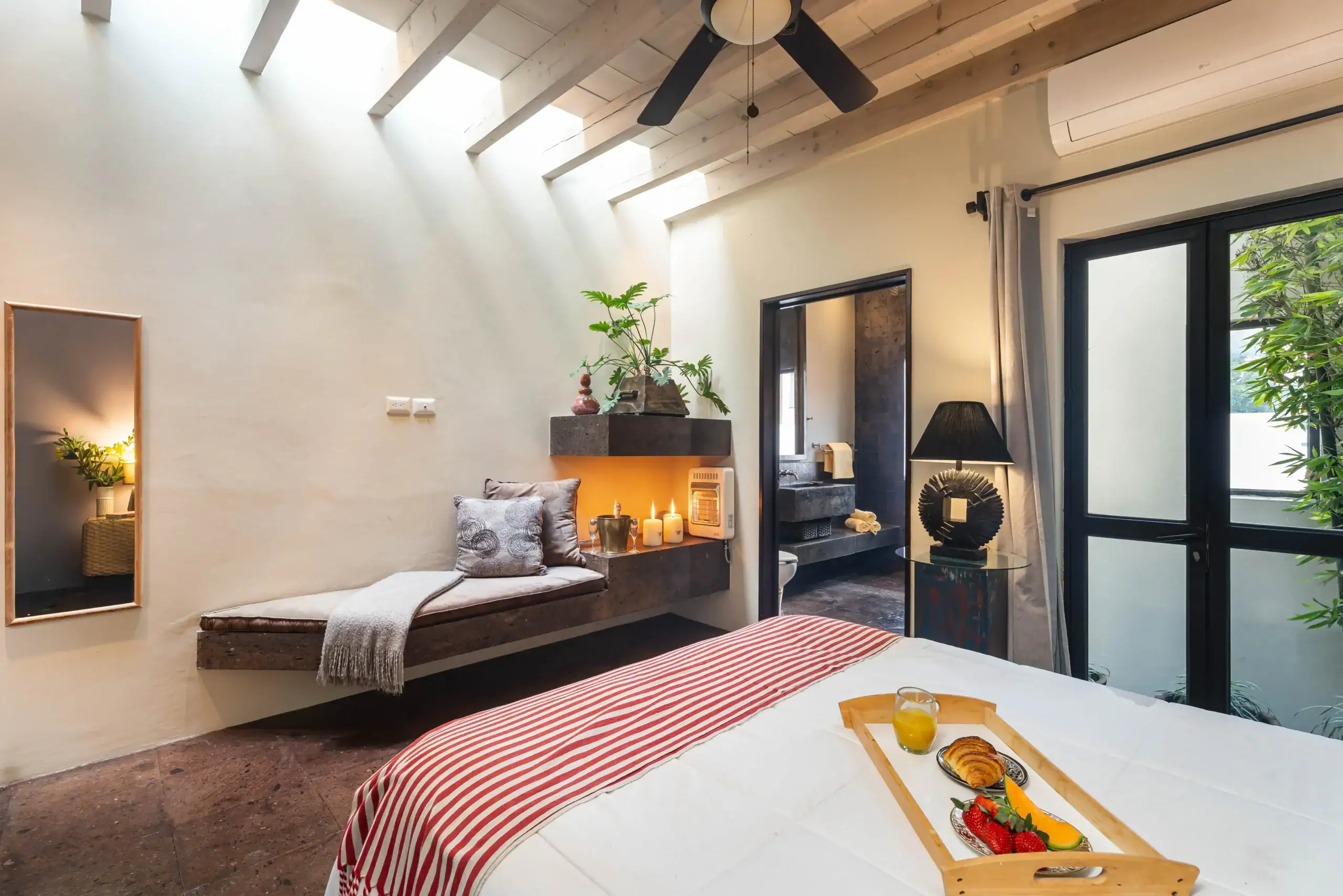 Breakfast tray with juice and pastry on a striped red-and-white bedspread in a bright, modern bedroom with skylight beams and plants nearby, San Miguel de Allende