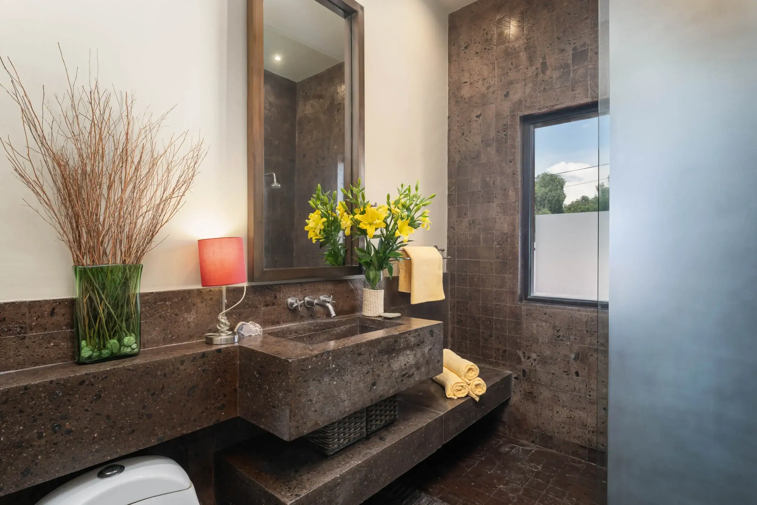 Bathroom vanity with dark brown marble countertop, vase of yellow lilies and dried branches, a red lamp, and a wall-mounted mirror above the sink, San Miguel de Allende