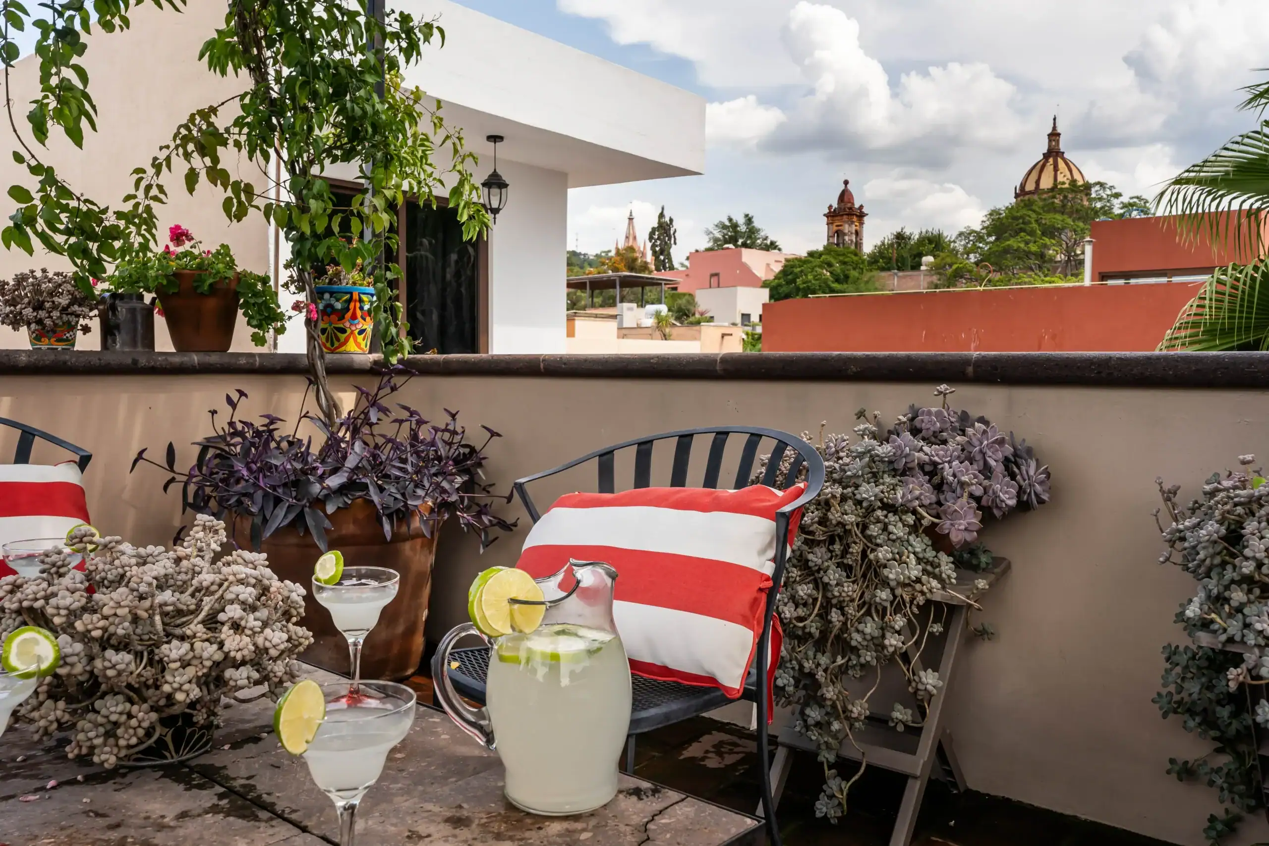 Outdoor terrace with a pitcher of lemonade and lime-edged cocktails on a table, red-and-white striped cushions, and potted plants against a white building and distant domed church, San Miguel de Allende