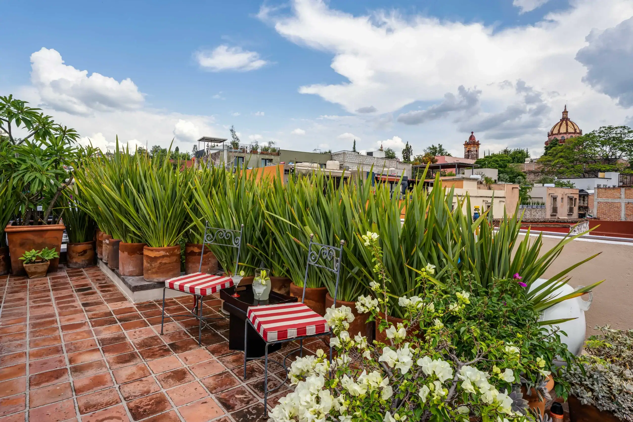 Rooftop terrace with terracotta tiles, potted plants, and red-and-white striped seating; city skyline with a domed church under a blue, partly cloudy sky, San Miguel de Allende