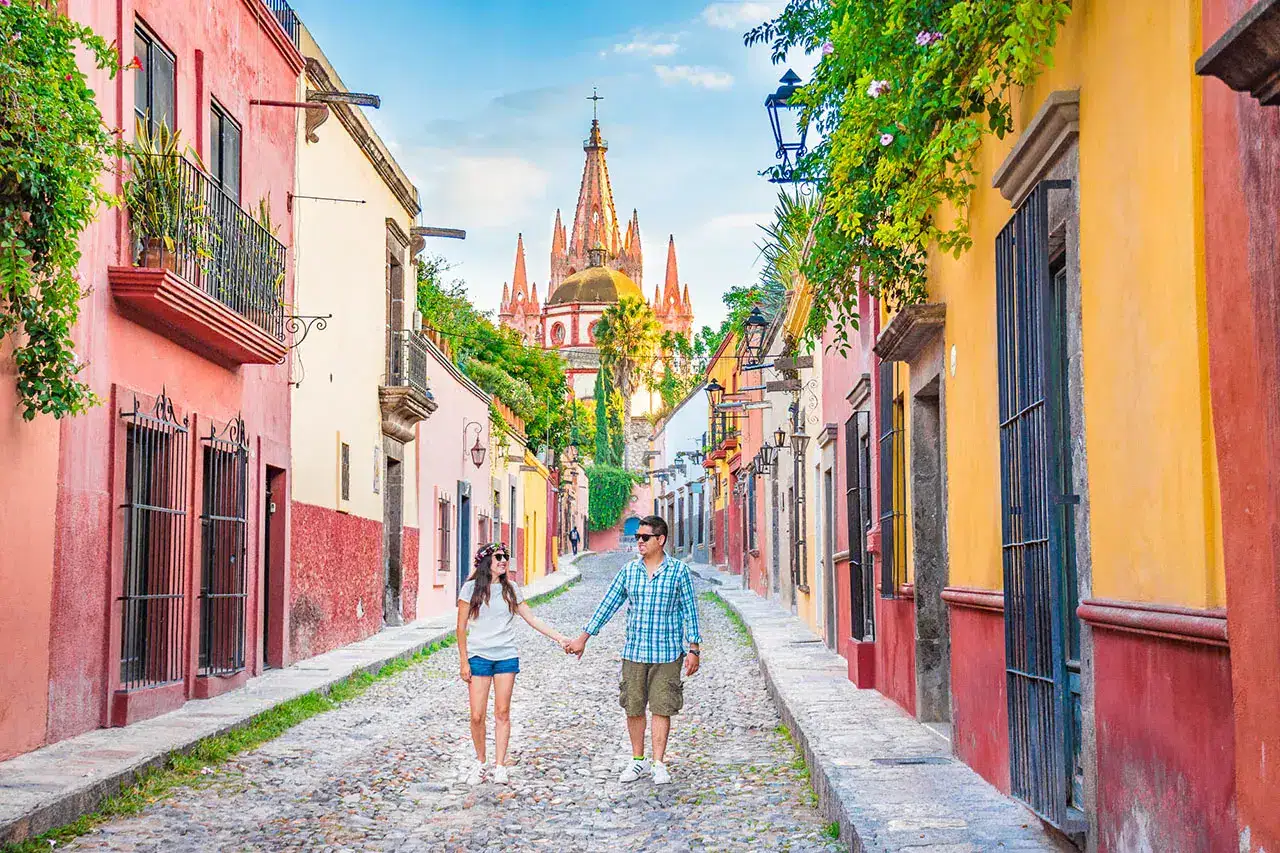 Couple holding hands and walking down a colorful cobblestone street with vibrant buildings and a church in the distance.