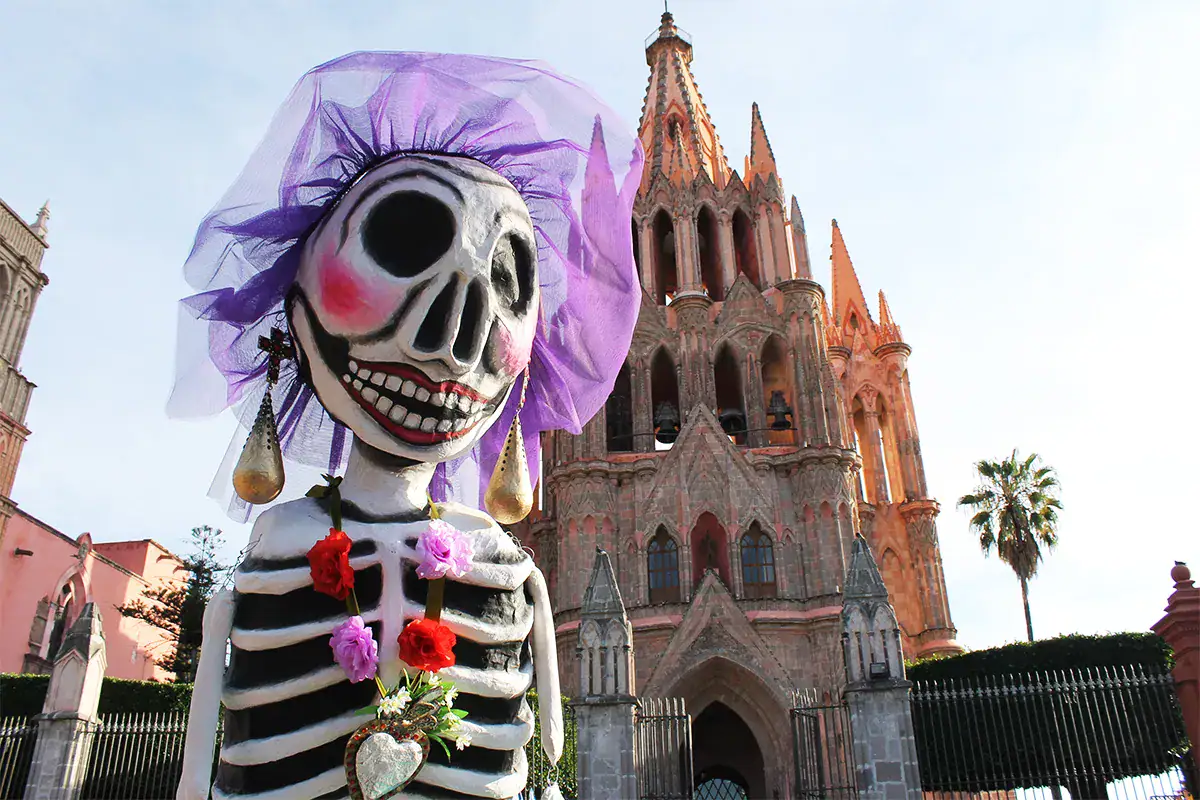 Oversized skeletal figure with a purple veil and flower garland in front of a pink Gothic church.