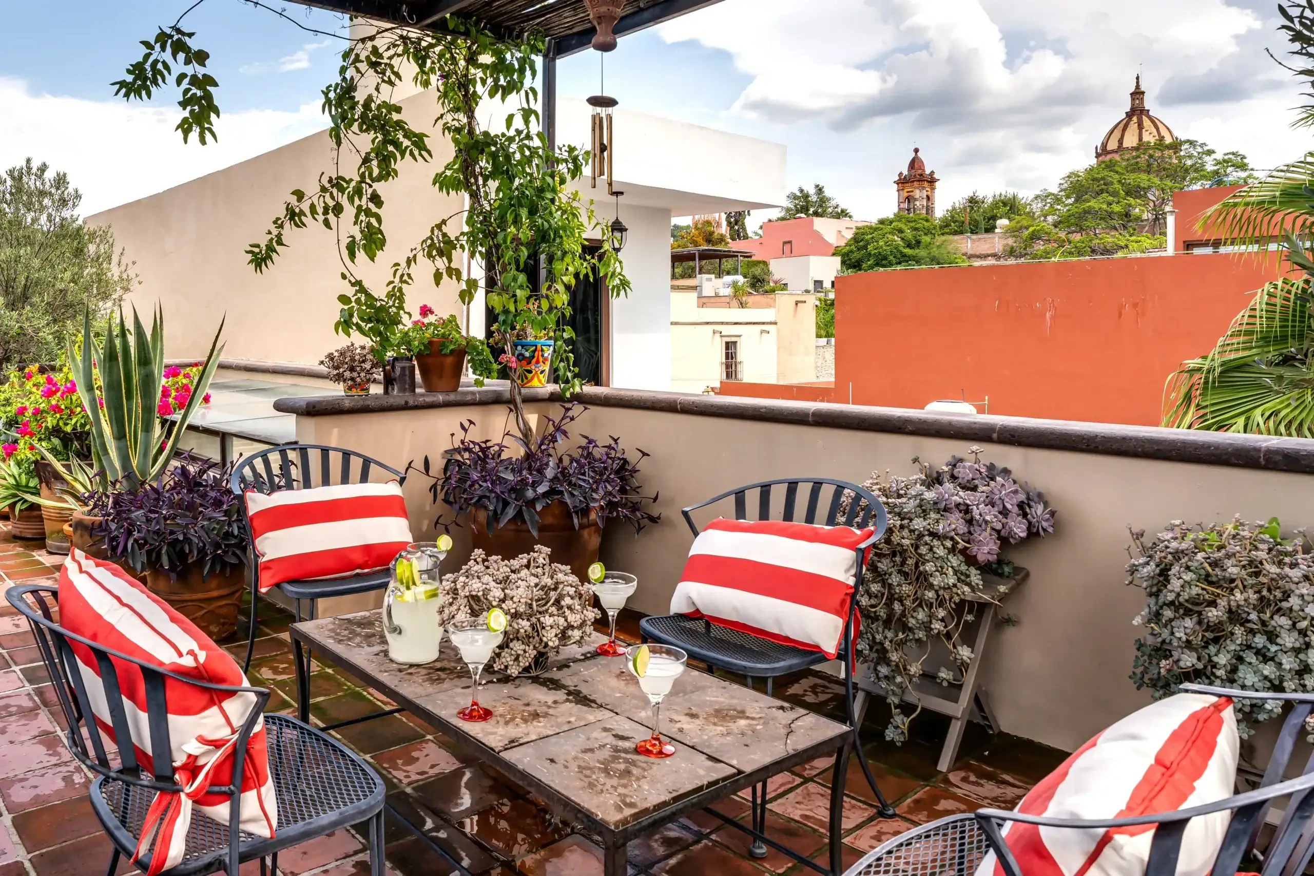 Rooftop patio with red-and-white striped cushions, metal chairs, and a table with drinks, surrounded by potted plants and a cityscape in the background, San Miguel de Allende