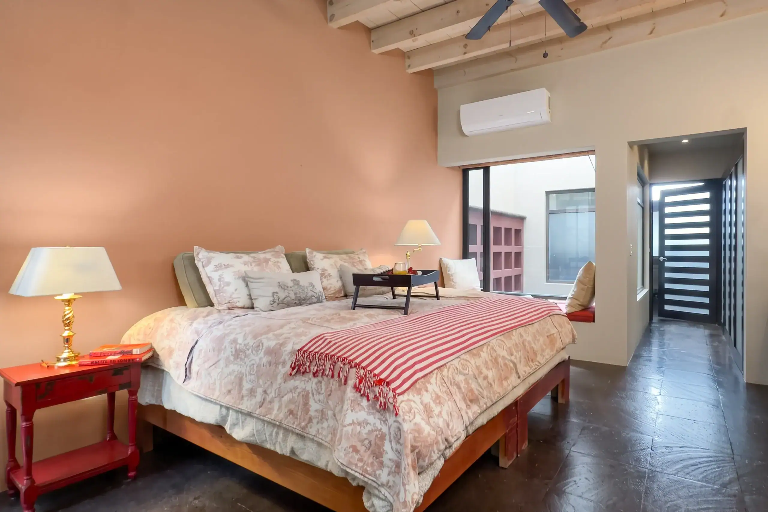 Bedroom with a pink accent wall, large bed with floral bedding, red nightstand, and warm lamps; exposed ceiling beams and a wall-mounted air conditioner, San Miguel de Allende