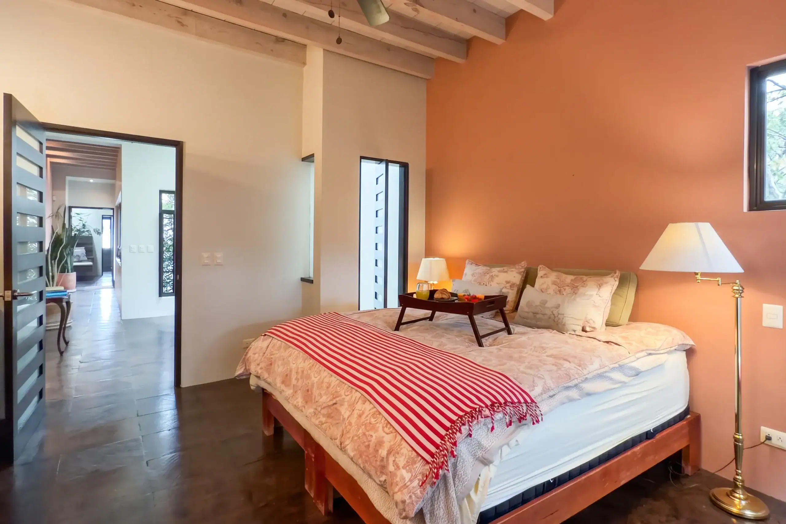 Bedroom with a queen bed, beige bedding and red-and-white striped blanket, two lamps, and an orange accent wall; doorway to a hallway on the left, San Miguel de Allende