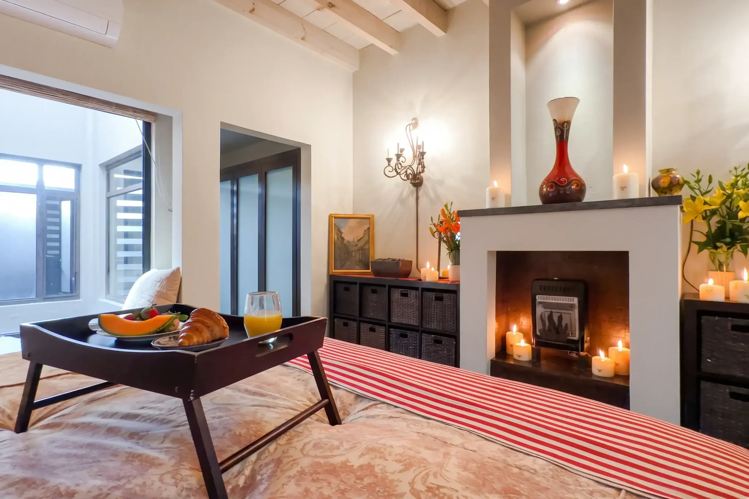 Cozy living room breakfast scene with a black tray holding a croissant, melon slices, and orange juice on a red-and-white striped rug by large windows, San Miguel de Allende