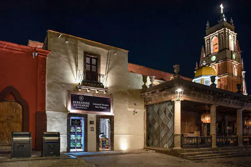 Night view of a white Colonial Homes storefront with a Berkshire Hathaway sign, next to a red wall and a lit church tower in the background.
